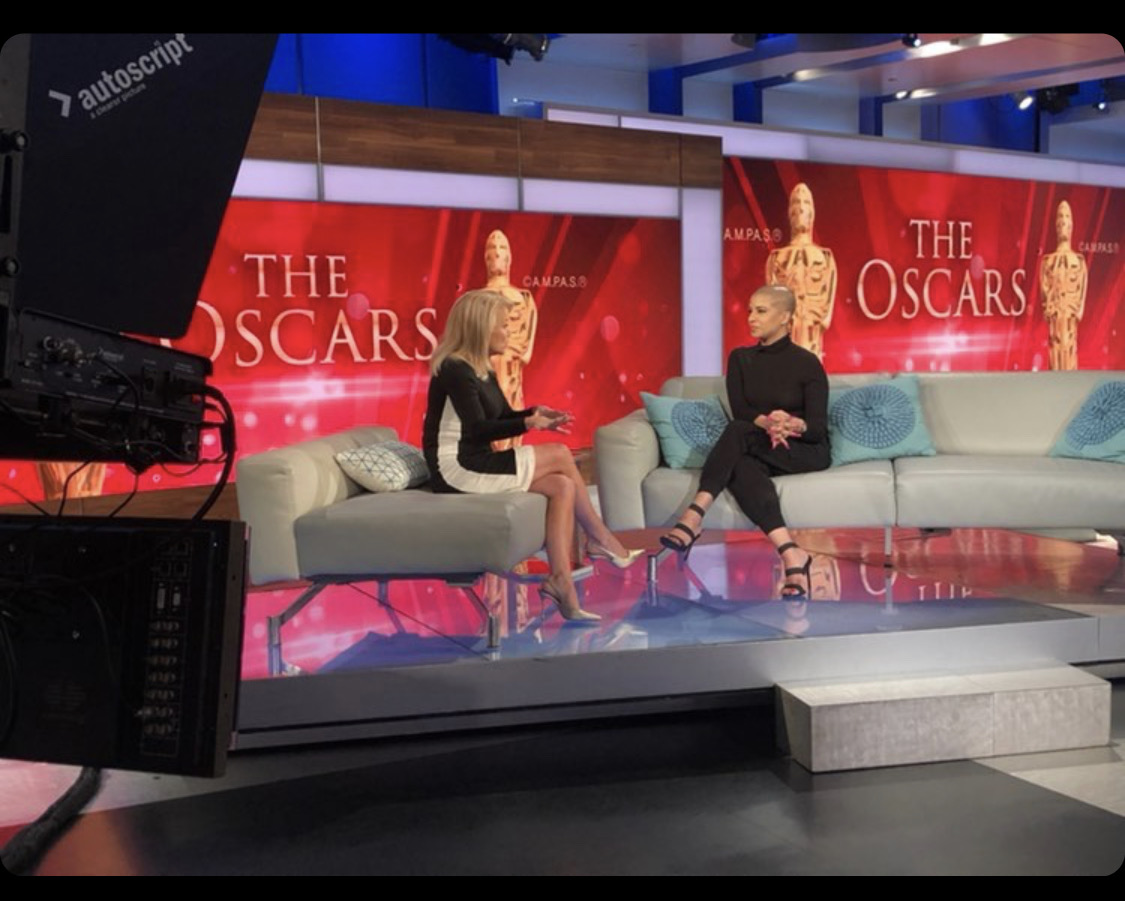Two women sit on a sofa in a TV studio with Oscar-themed background, discussing during a broadcast.
