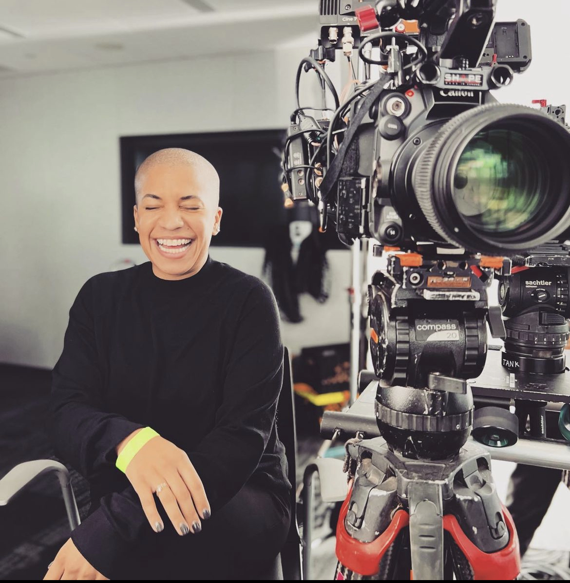Bald woman laughing, sitting in a room with a large camera on a tripod nearby, smiling with eyes closed, wearing a black top.