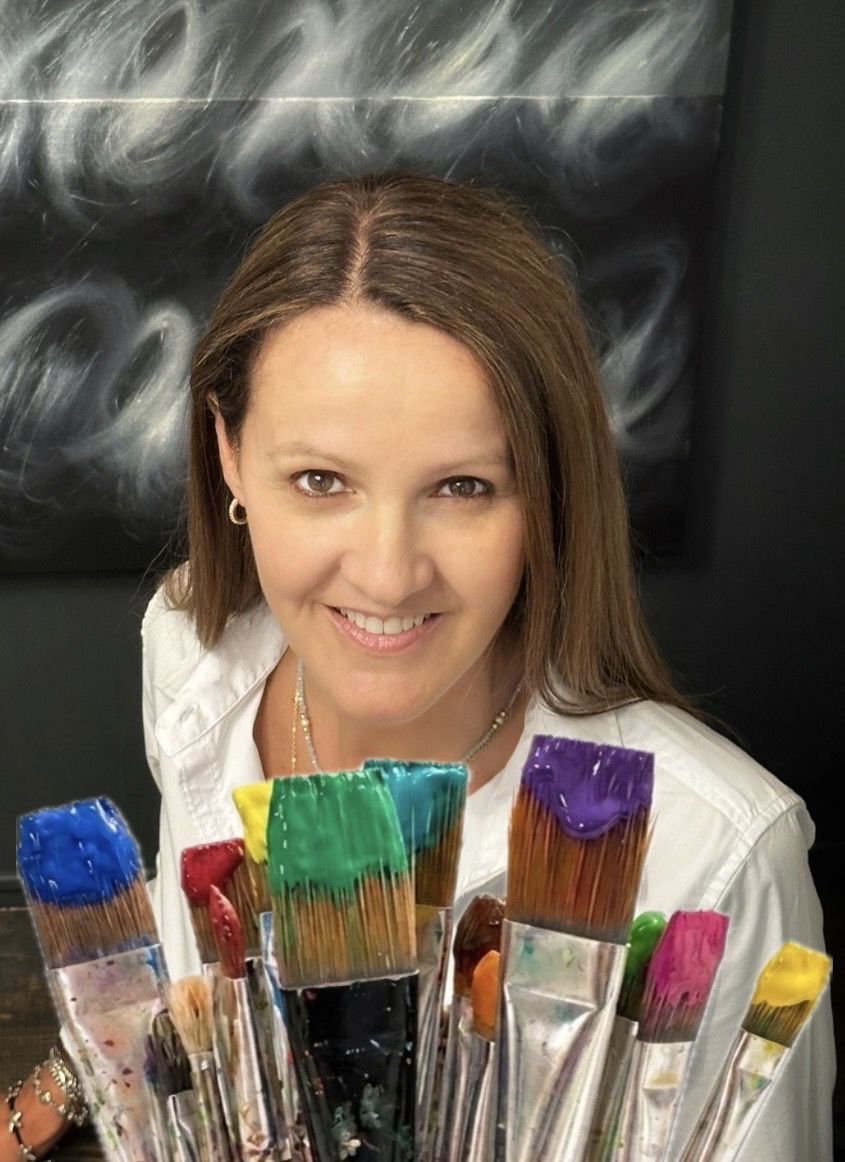 Woman with brown hair smiling, surrounded by colorful paintbrushes, in front of a blackboard with chalk marks.
