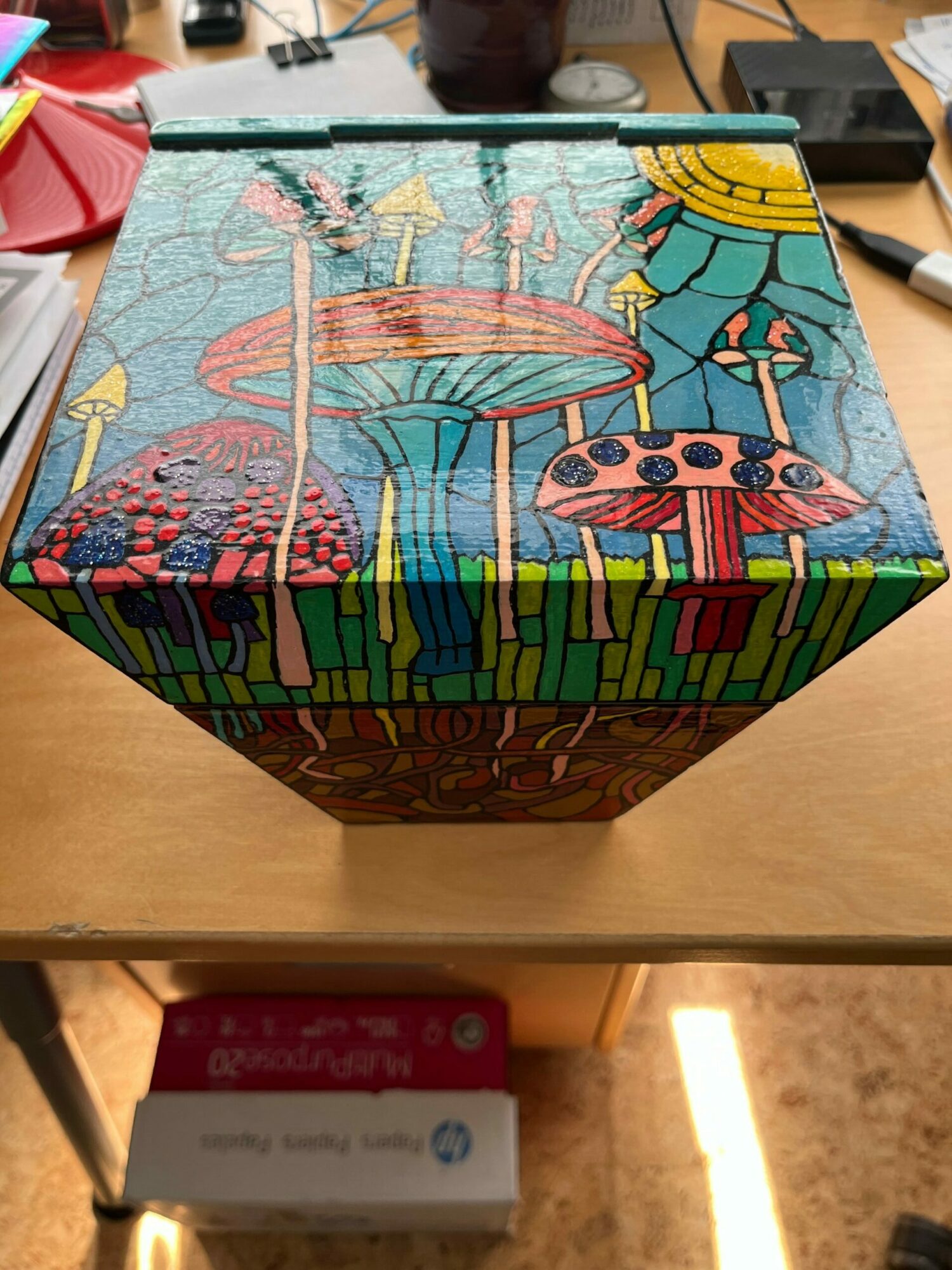 Colorful, illustrated box with a landscape scene, including a fountain, trees, and a sun, on a wooden surface.