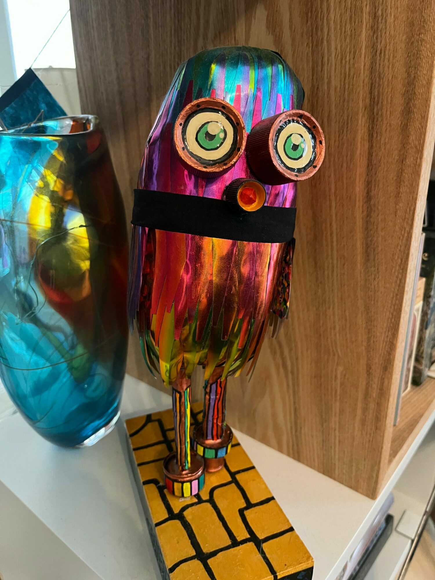 Colorful robot sculpture with large eyes, a black band, and a yellow brick wall pattern on the back, placed on a white surface.