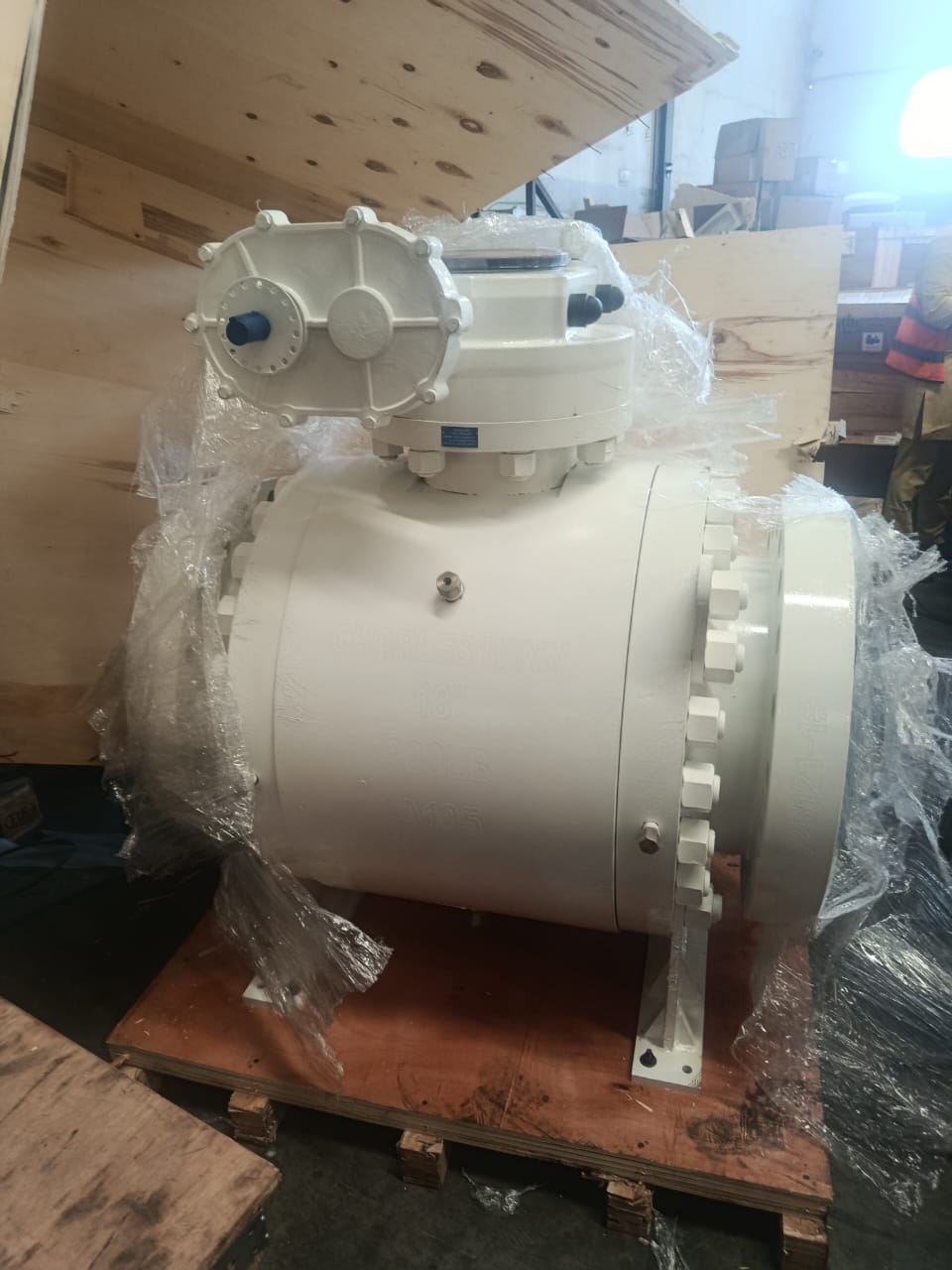 Large white industrial valve with multiple bolts, wrapped in plastic, on a wooden pallet in a workshop.