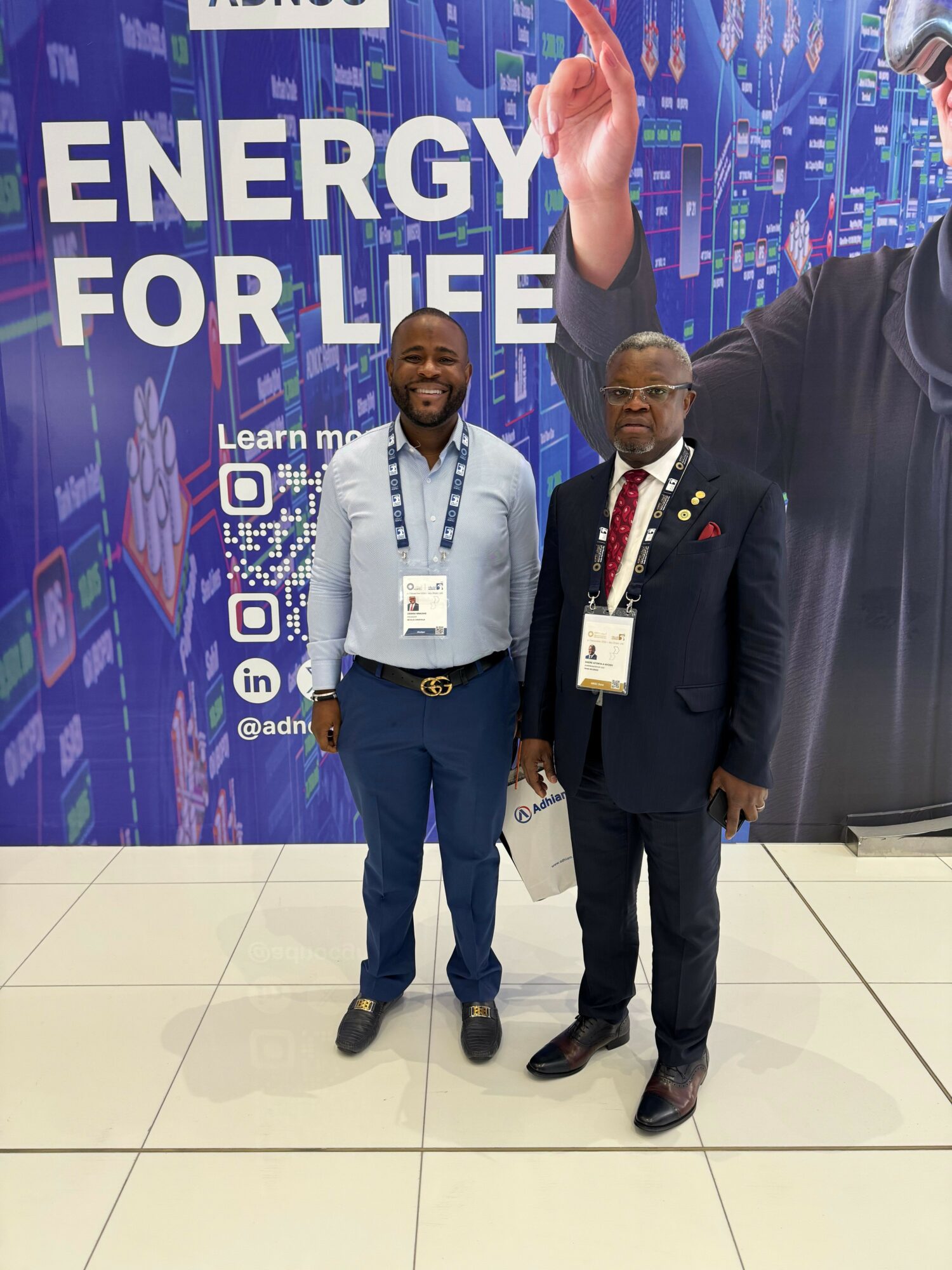 Two people standing in front of a blue backdrop with text, one in a suit and the other in casual attire, both wearing conference badges.