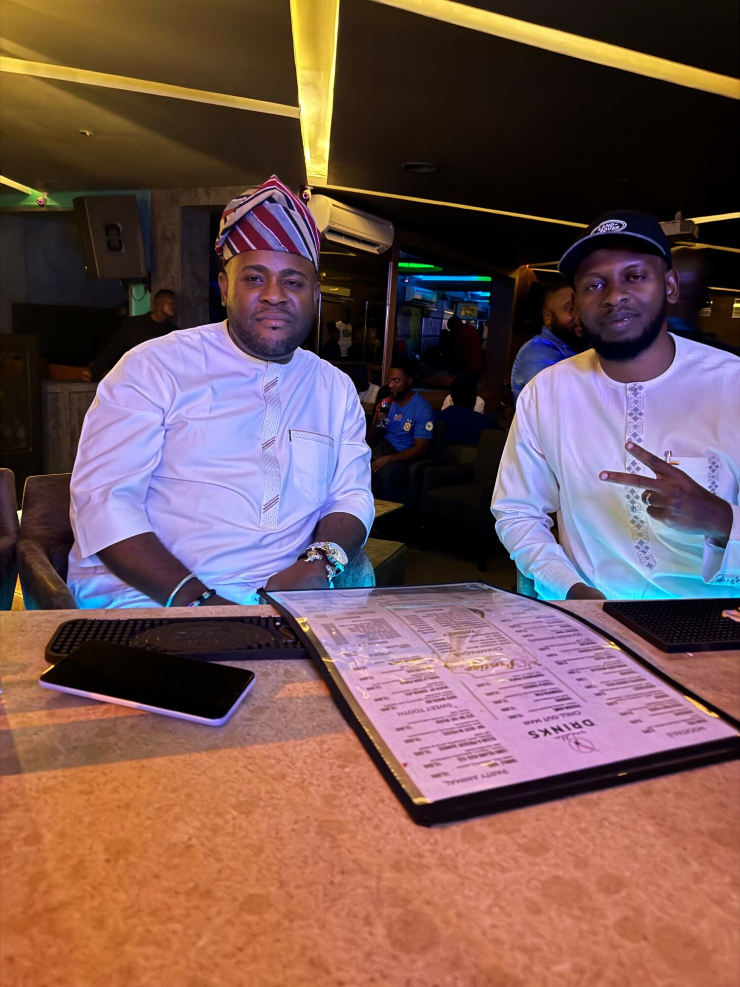 Two men sit at a table in a dimly lit restaurant, one looking at a menu, the other pointing at it.