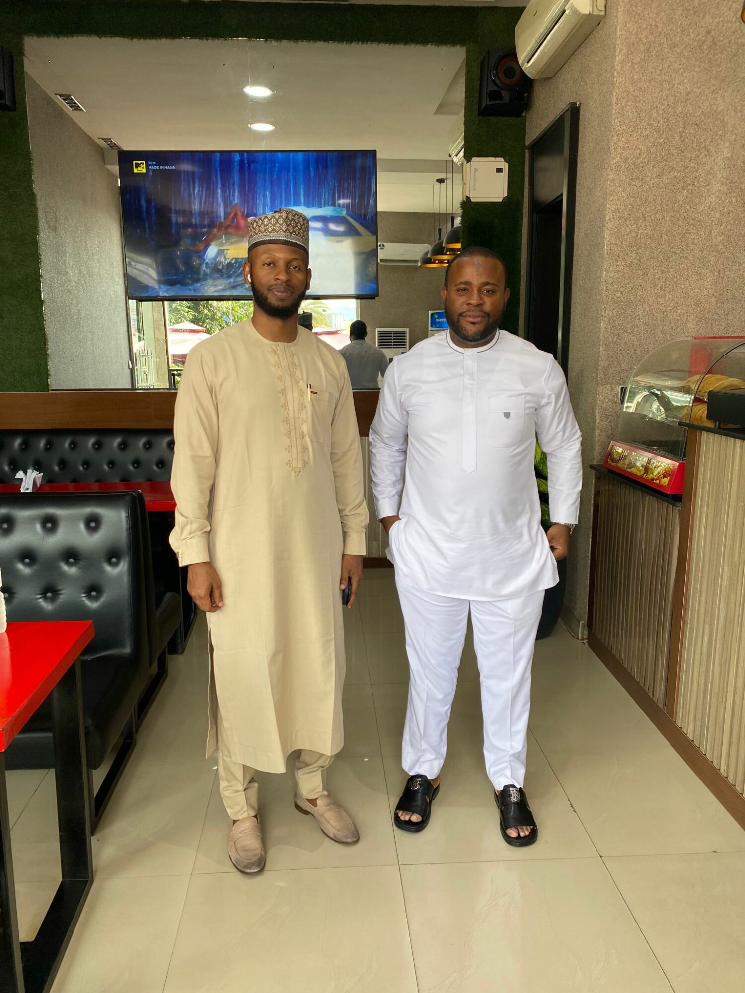 Two men stand side by side indoors, one in beige traditional attire and the other in white outfit, with a TV and seating area behind them.