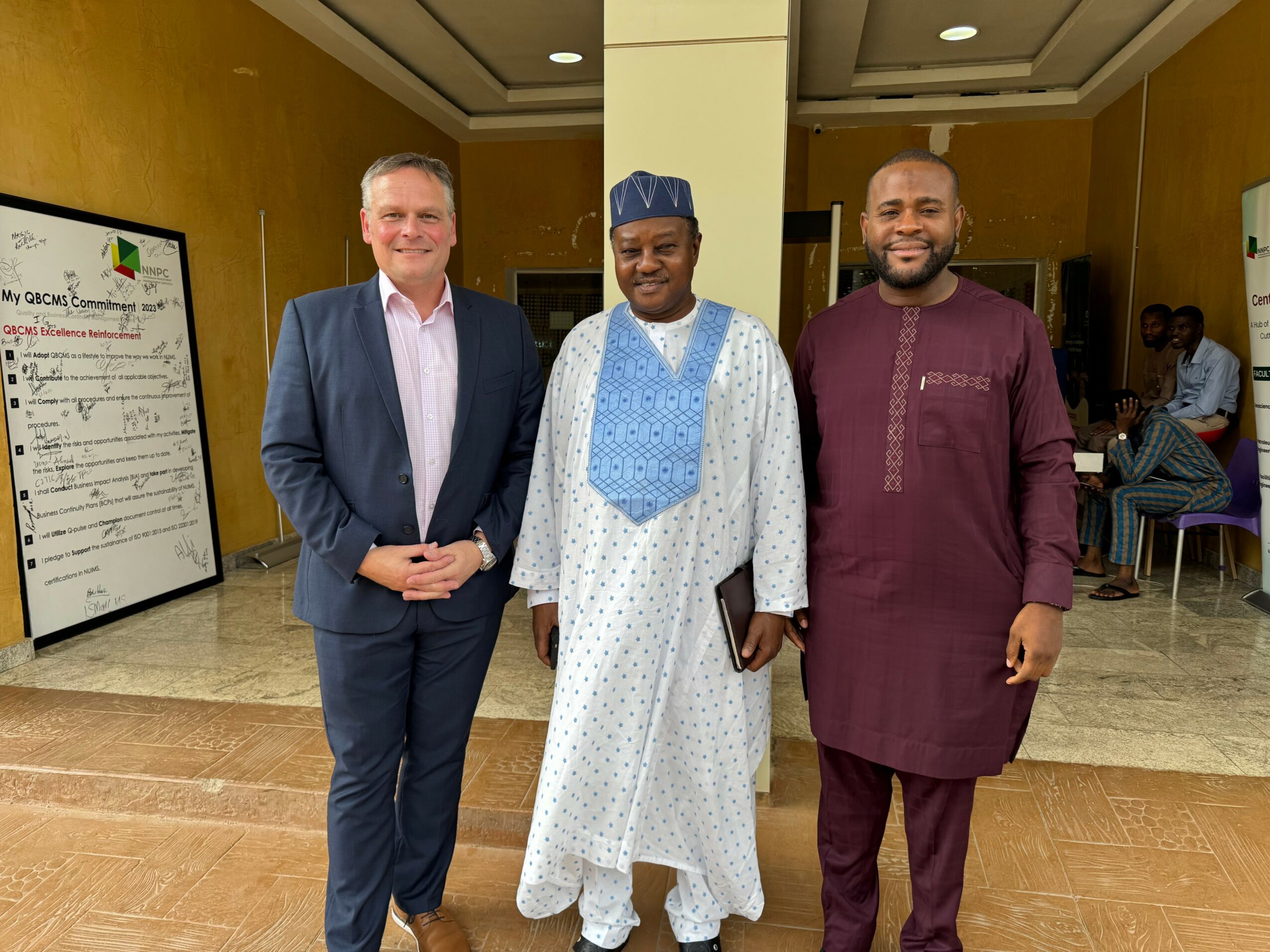 Three men standing indoors, two in traditional attire and one in a suit, smiling at the camera.