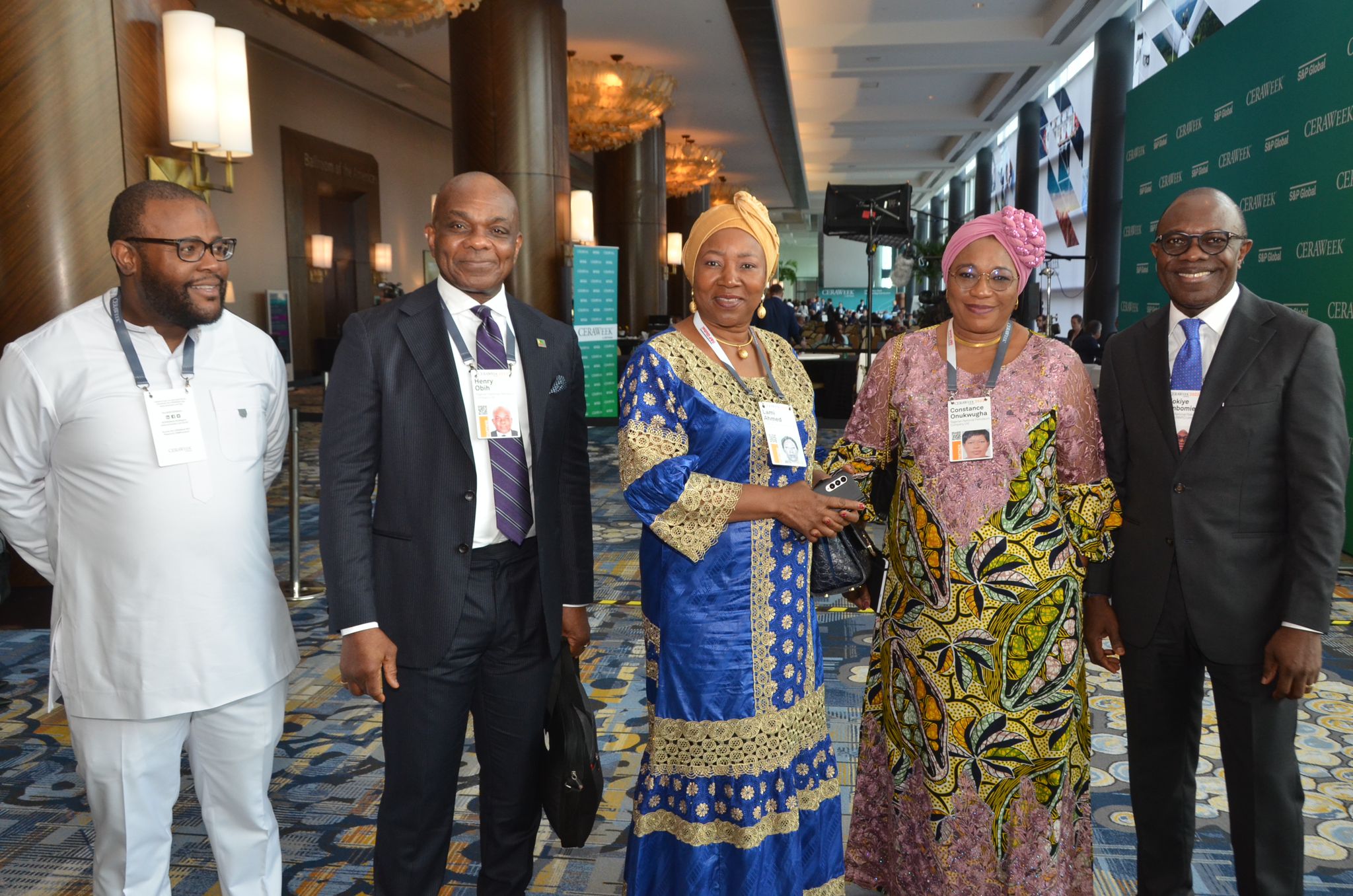 Group of five people standing together indoors, smiling, dressed in formal and traditional attire, with a patterned carpet and background decor.