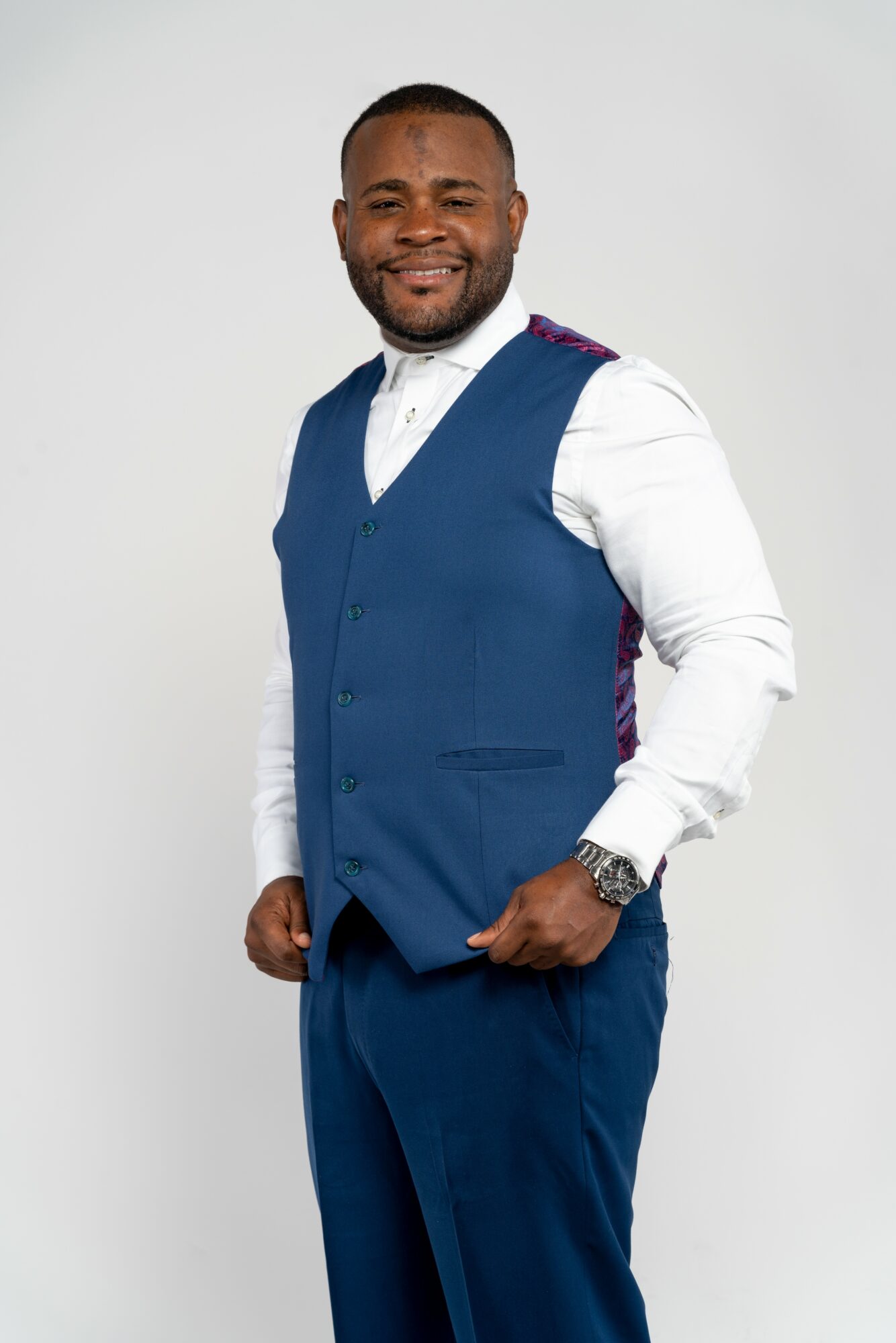 Smiling man in a blue vest and pants, white shirt, standing with hands in pockets against a plain background.