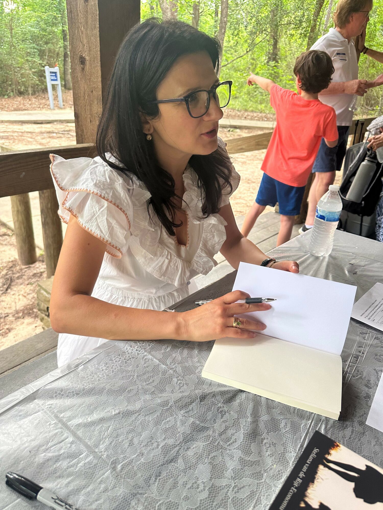 Woman with glasses and dark hair sitting at a table outdoors, writing in a notebook, with children in background.