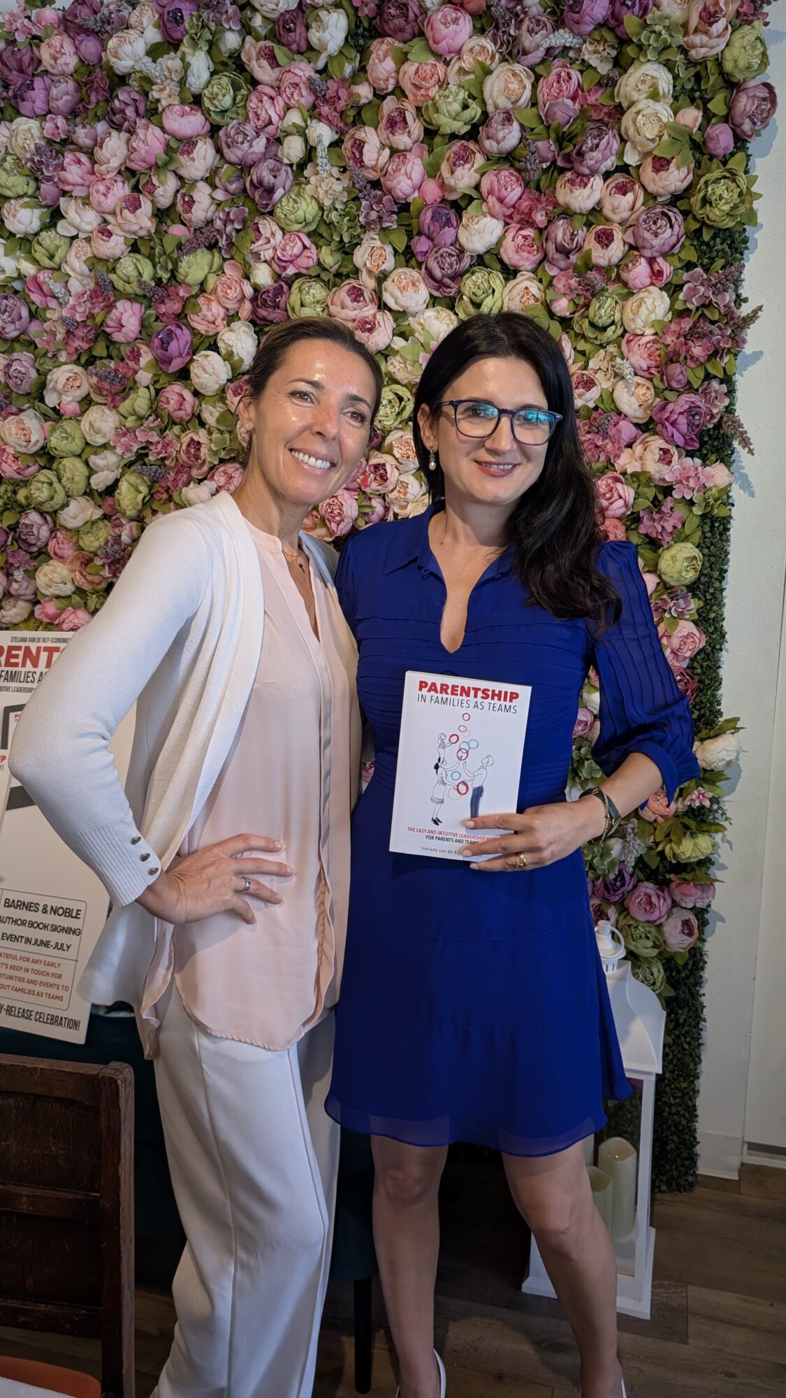 Two women standing in front of a floral wall, one holding a certificate, smiling at the camera.