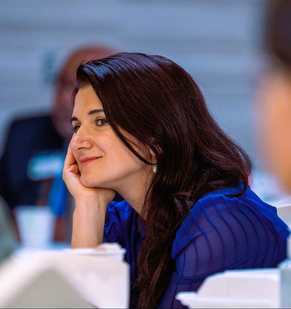 Woman with dark hair smiling, resting her chin on her hand, wearing a blue top, in a meeting setting.