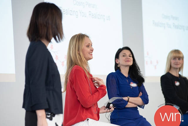 Four women standing and speaking on stage during a presentation or panel discussion.