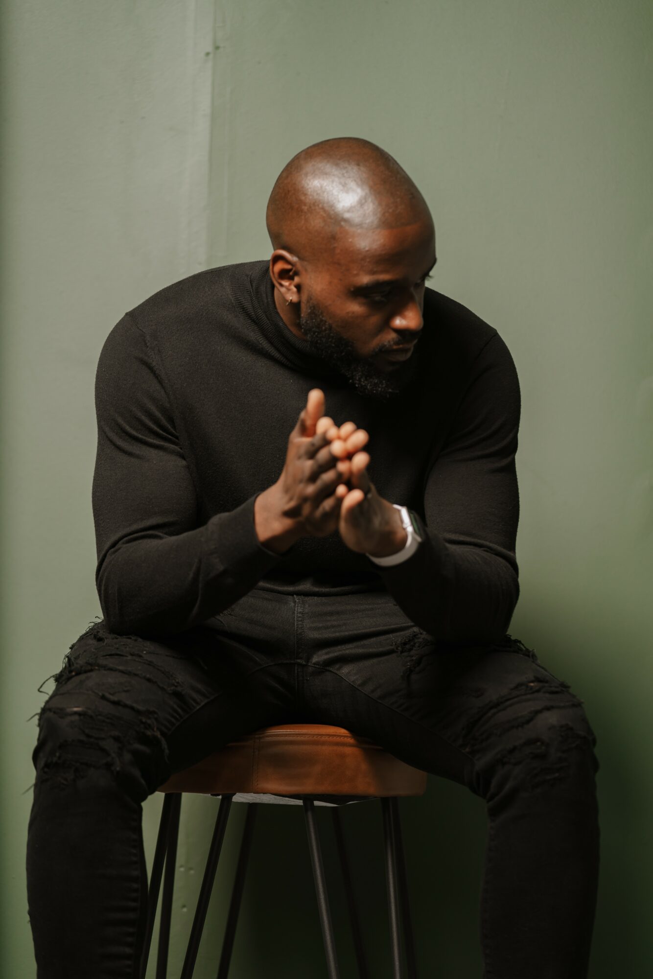 Man with a shaved head and dark clothing sitting on a chair, hands clasped, looking down, against a green wall.
