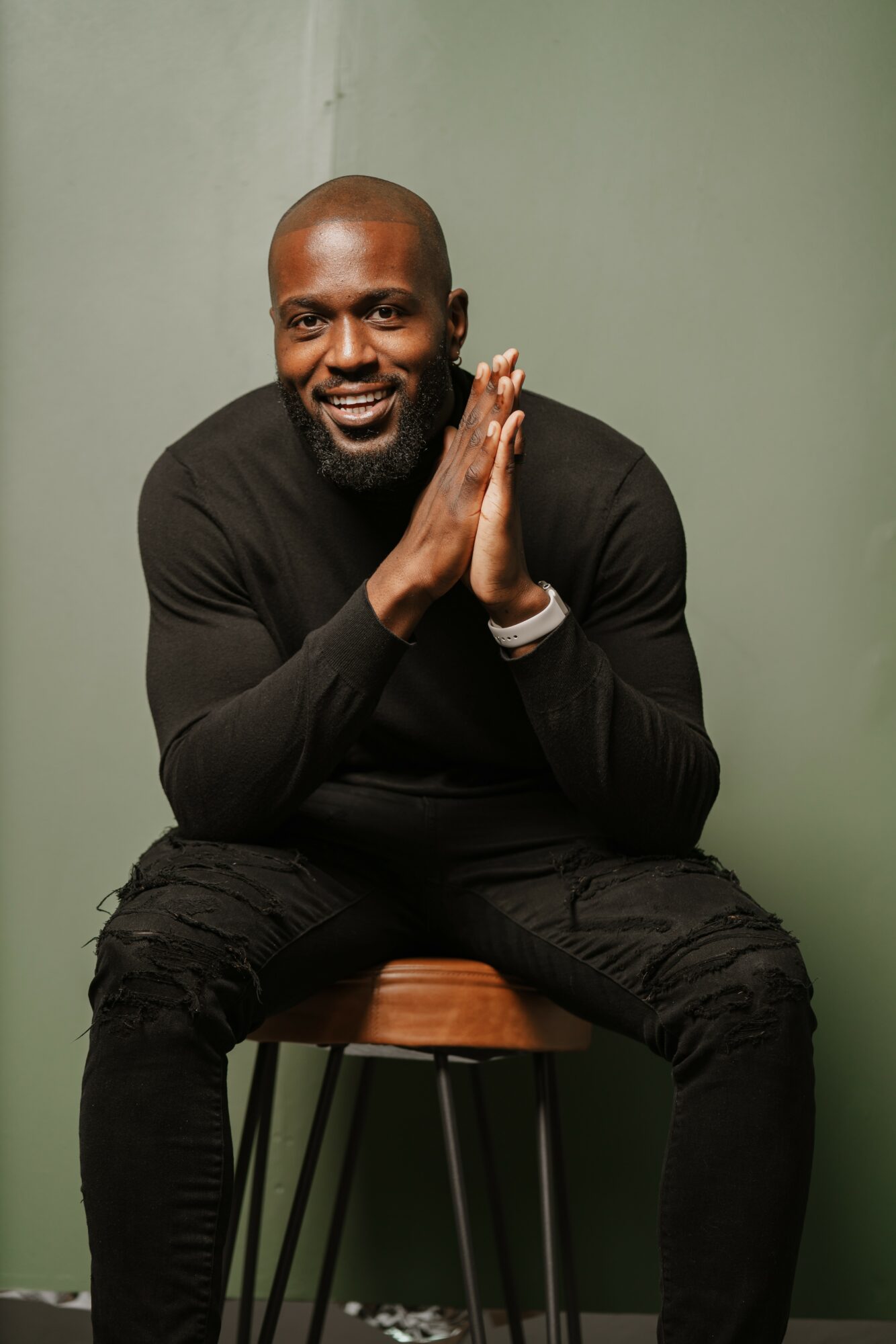 Smiling man with a beard, sitting on a stool, hands together, wearing a black long-sleeve shirt and pants, against a green wall.