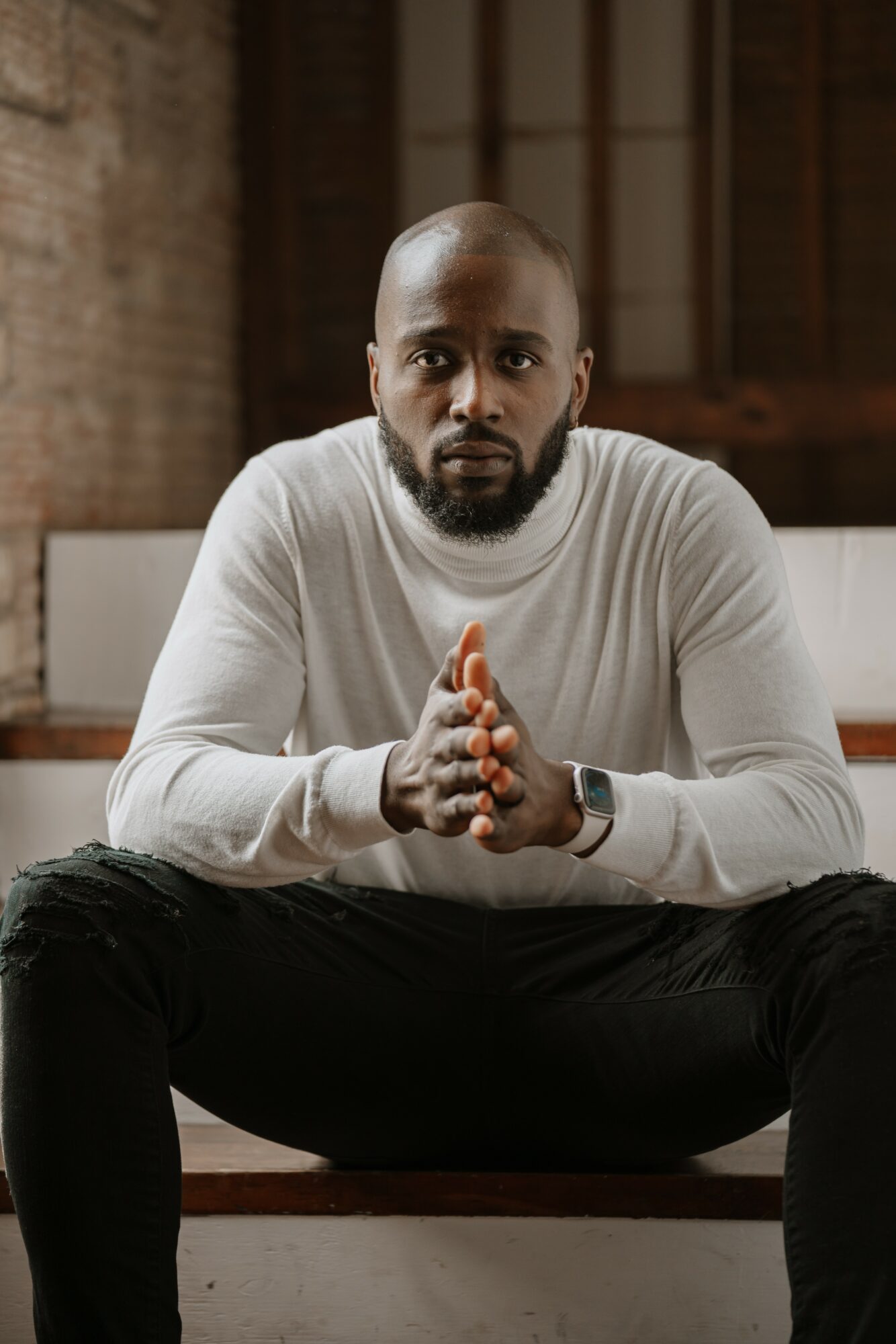 Man with a serious expression, sitting with hands clasped, wearing a white long-sleeve shirt, in an industrial-style room.