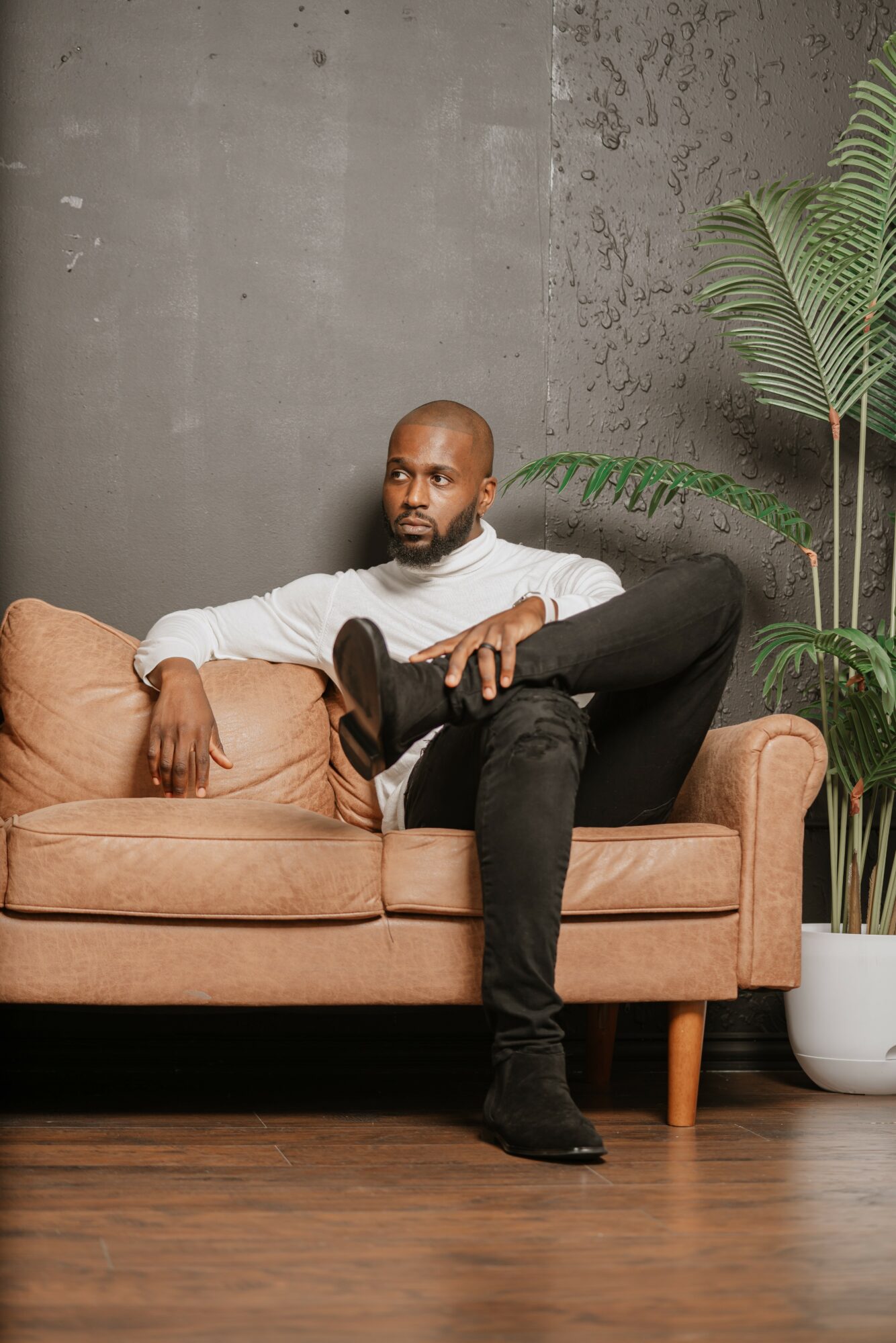 Man sitting on a sofa with one leg crossed over the other, next to a large potted plant, against a textured wall.