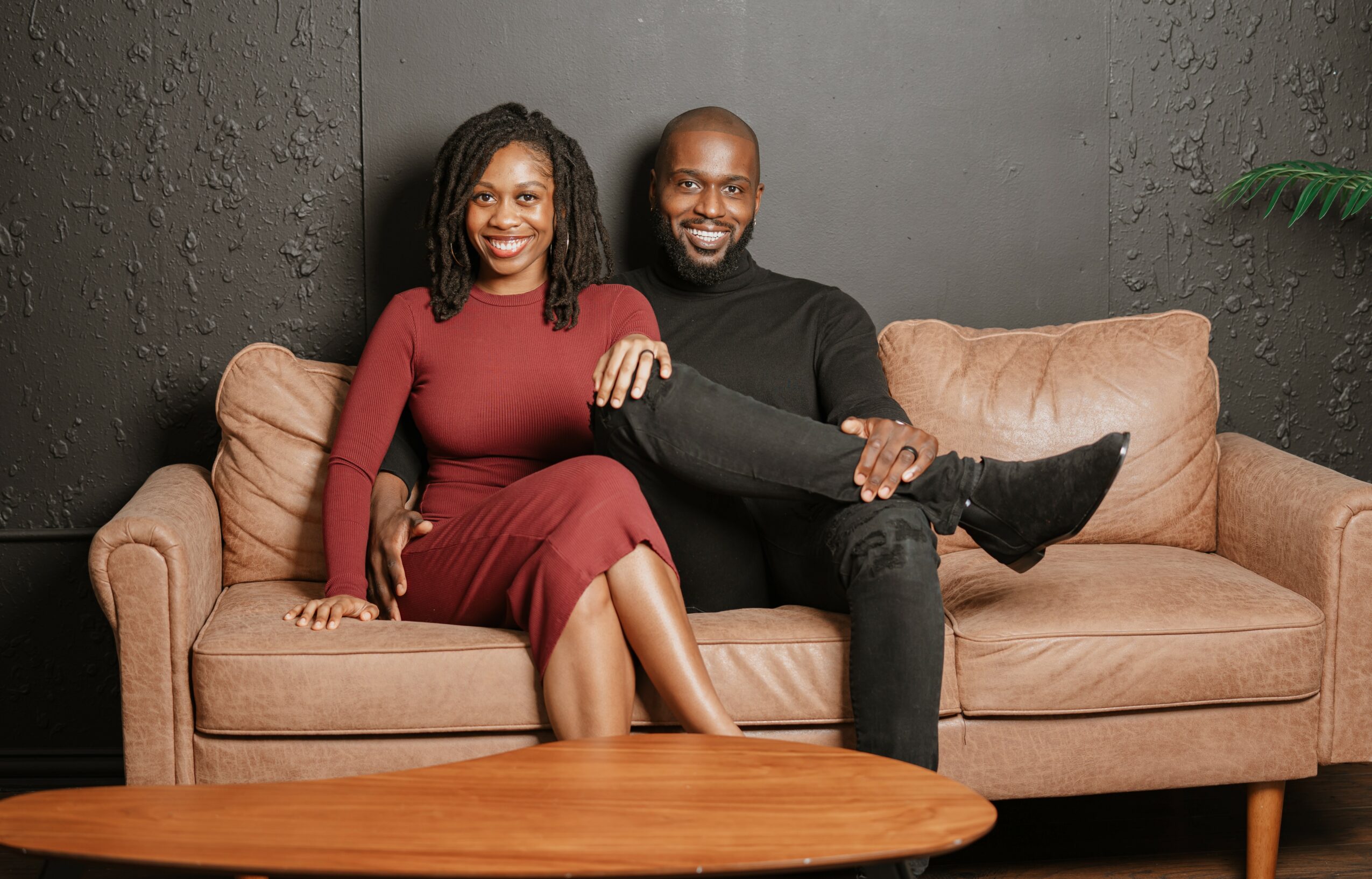 Two people sitting on a beige sofa, smiling, with a dark wall background and a wooden table in front.