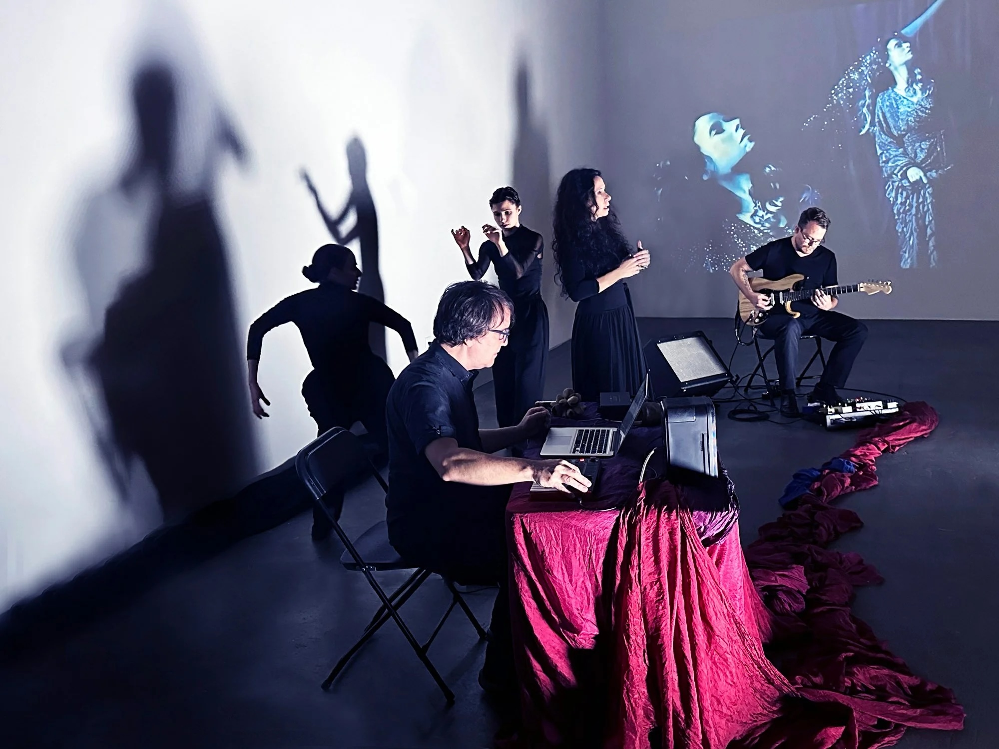 Group of musicians performing with shadows cast on the wall, some playing instruments and others singing, with a large screen behind them.