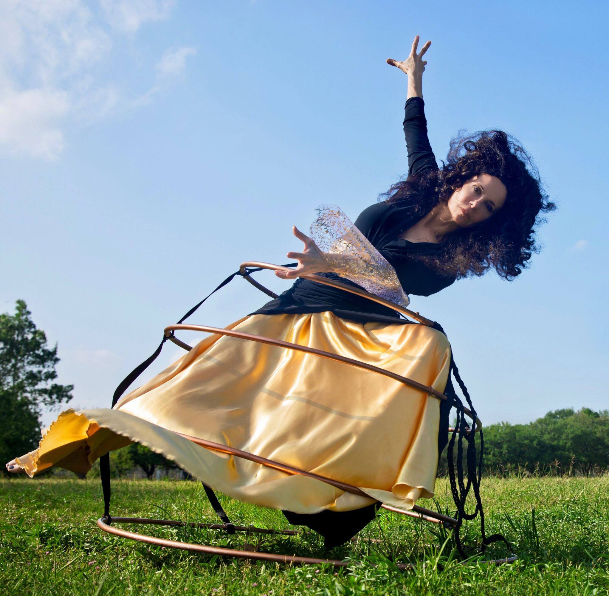 Person with curly hair in a black top and gold skirt appears to be falling or floating outdoors, with a blue sky and green landscape.