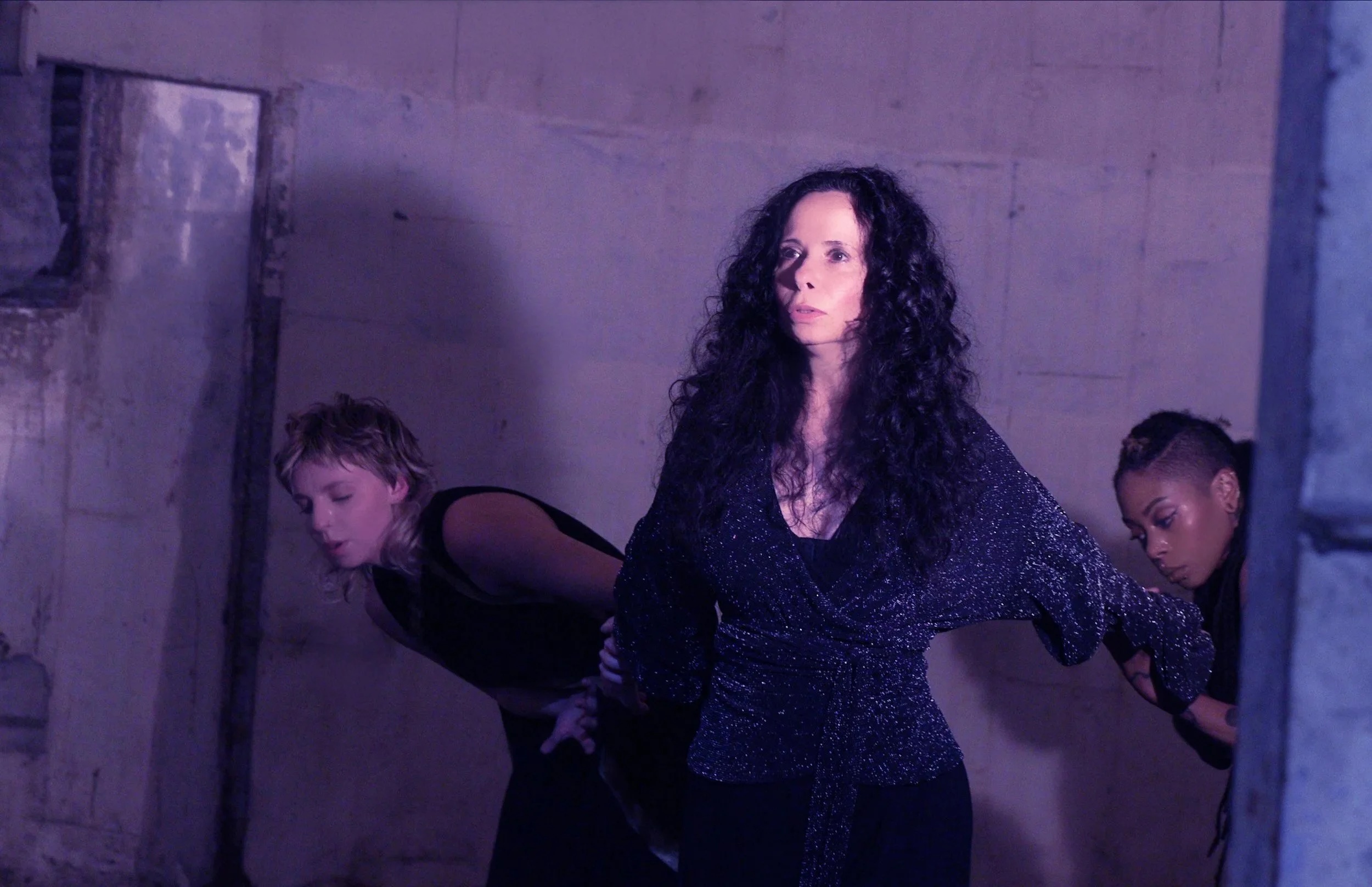 Three women in dark clothing pose against a concrete wall, with one woman in the center and two behind her.