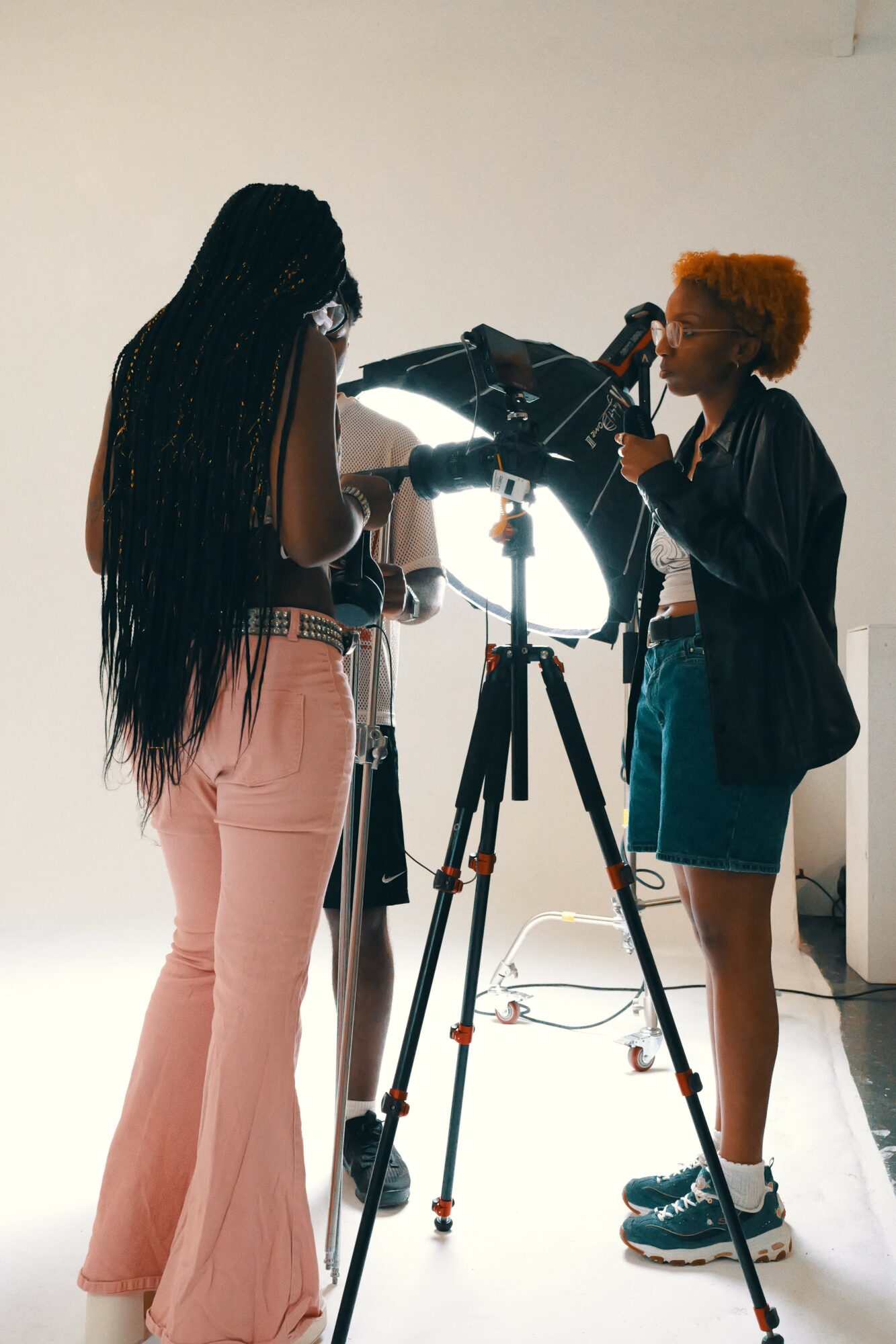Two women with a camera on a tripod, one with long braided hair and the other with short curly hair, in a studio.