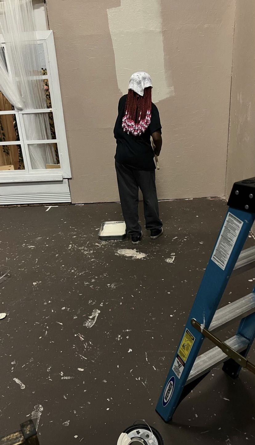 Person with pink and white braided hair, wearing a white cap, black shirt, and dark pants, standing against a beige wall.