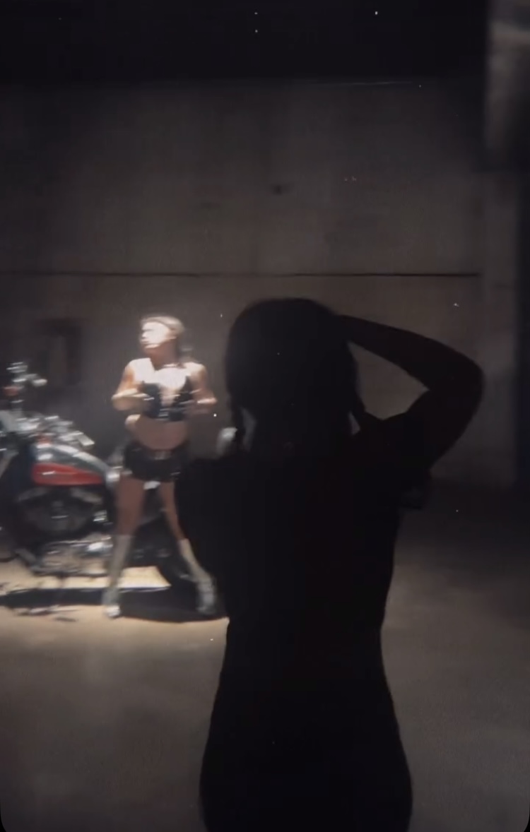 Silhouette of a person with hand on head, background shows a woman and motorcycles, dim lighting, concrete wall.
