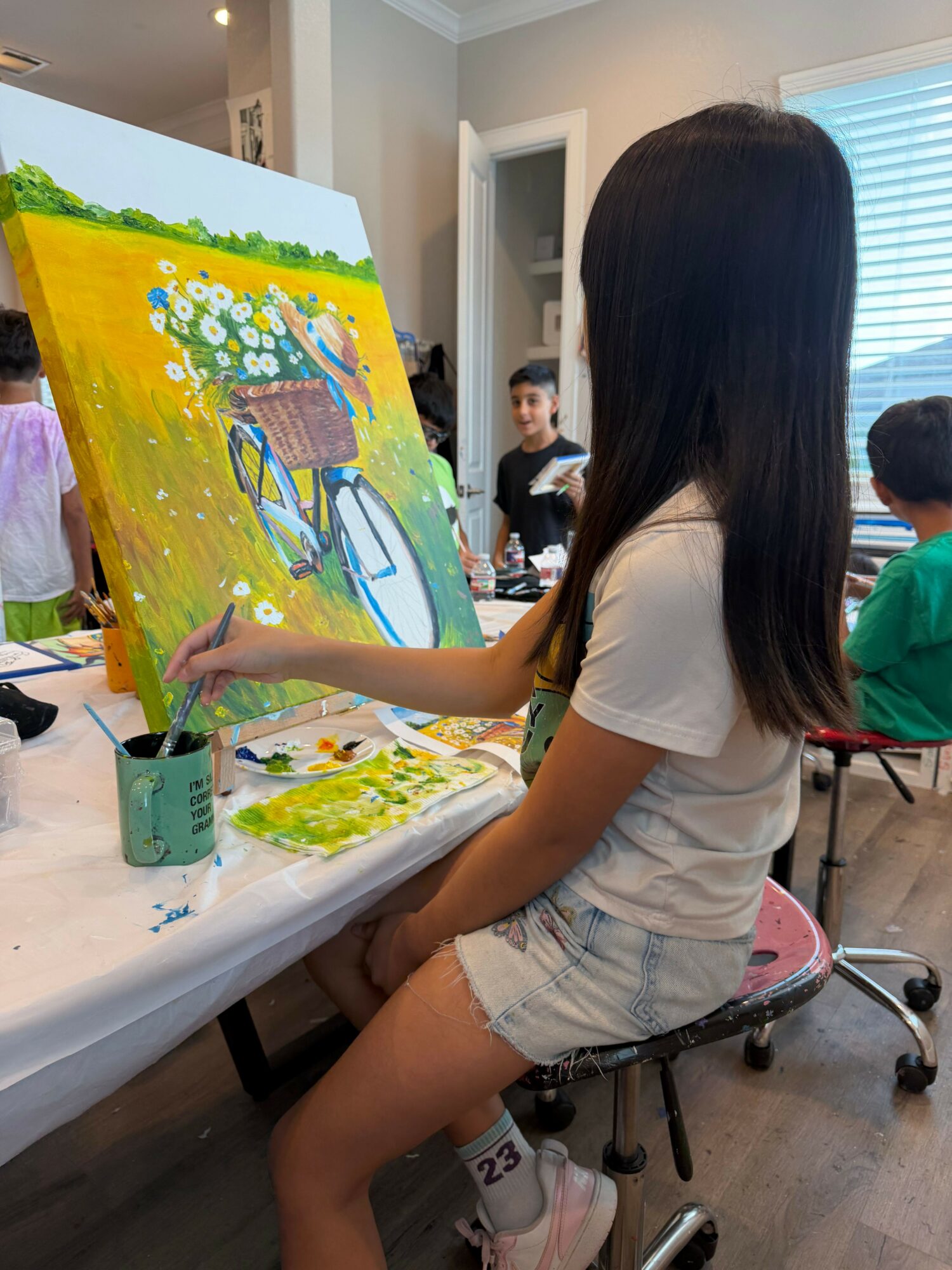 Girl painting a colorful outdoor scene on a canvas, with people and a building in the background.