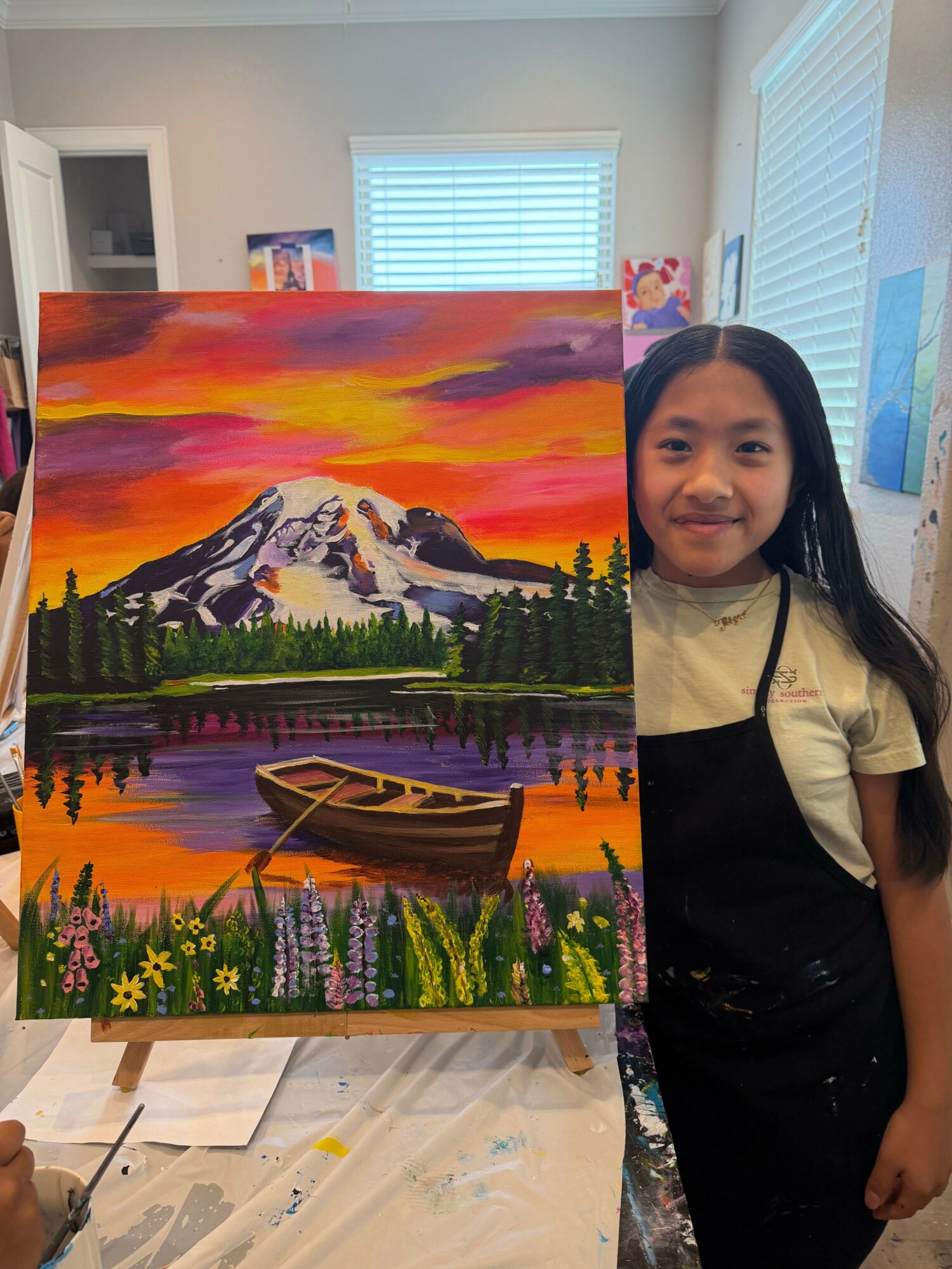Woman standing next to a colorful landscape painting of mountains, lake, trees, and a sunset sky.