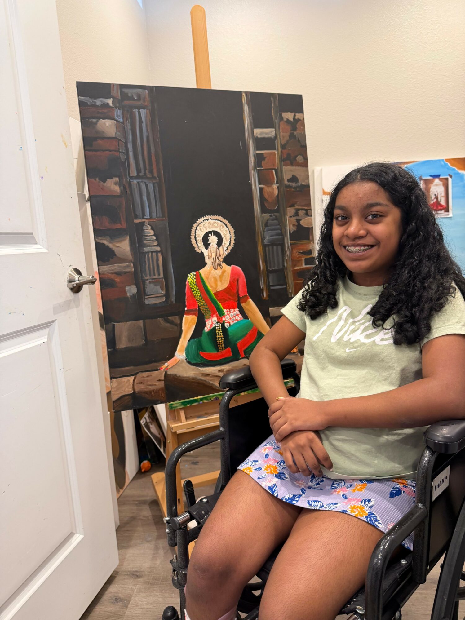 Young woman with curly hair sitting on a chair, wearing a light green top and patterned shorts, in a room with a fireplace and decorations.