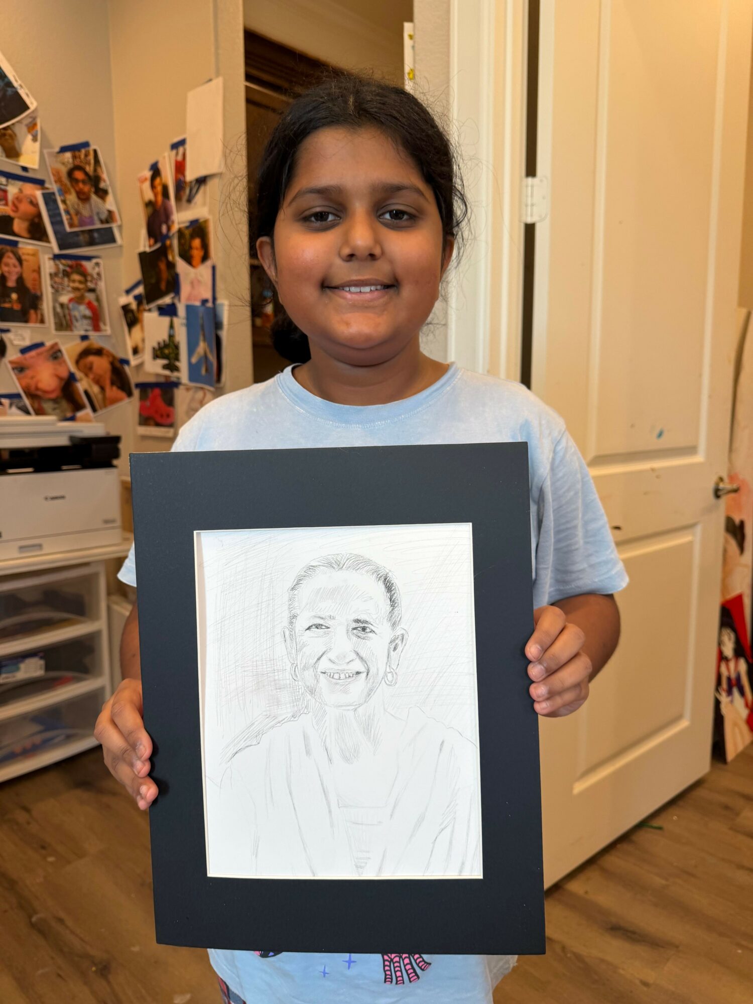 Girl holding a framed pencil sketch of a person's face, smiling, in a room with photos on the wall.