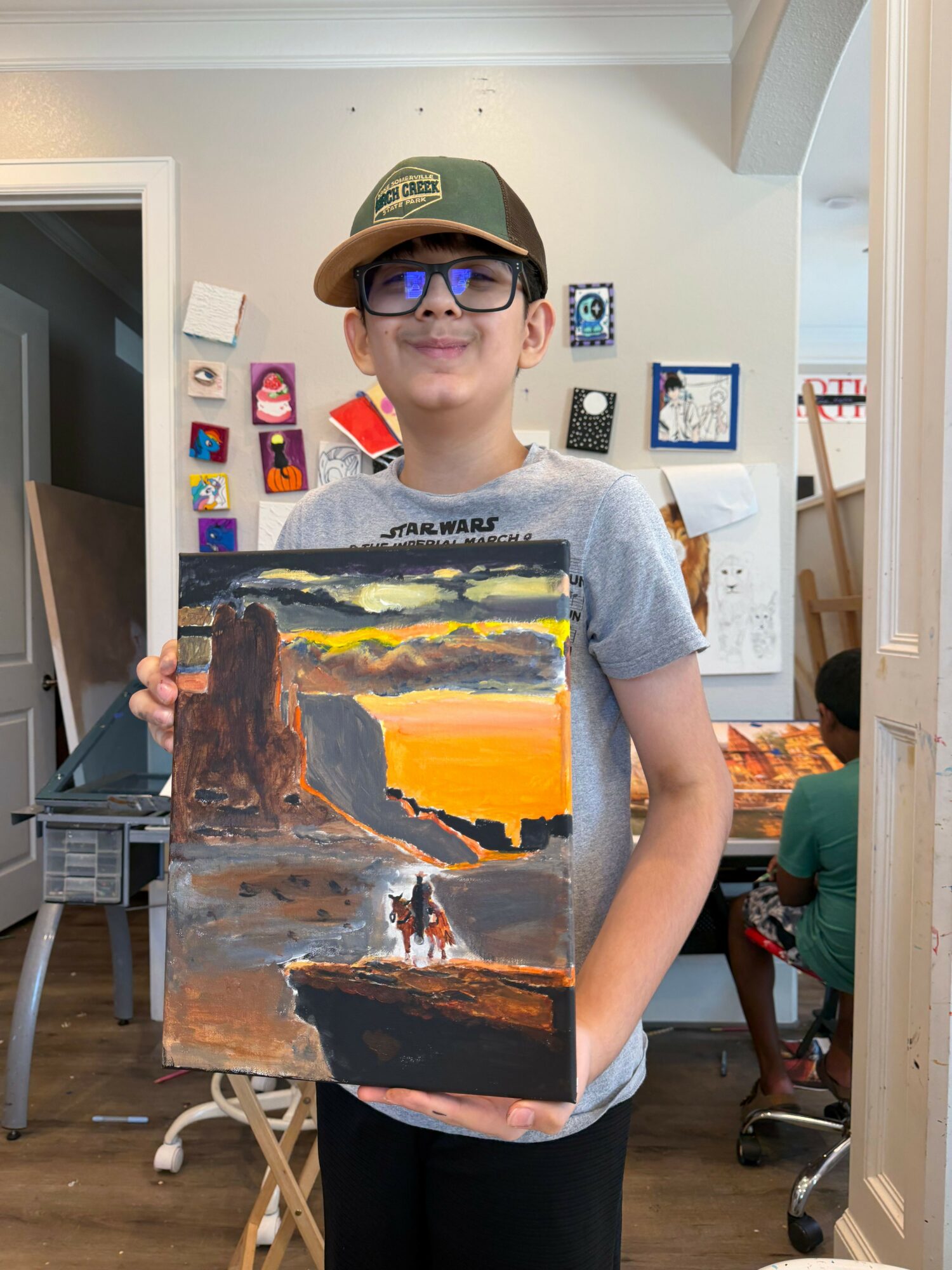 Person with glasses and a hat holding a landscape painting in a room with artwork on the wall.