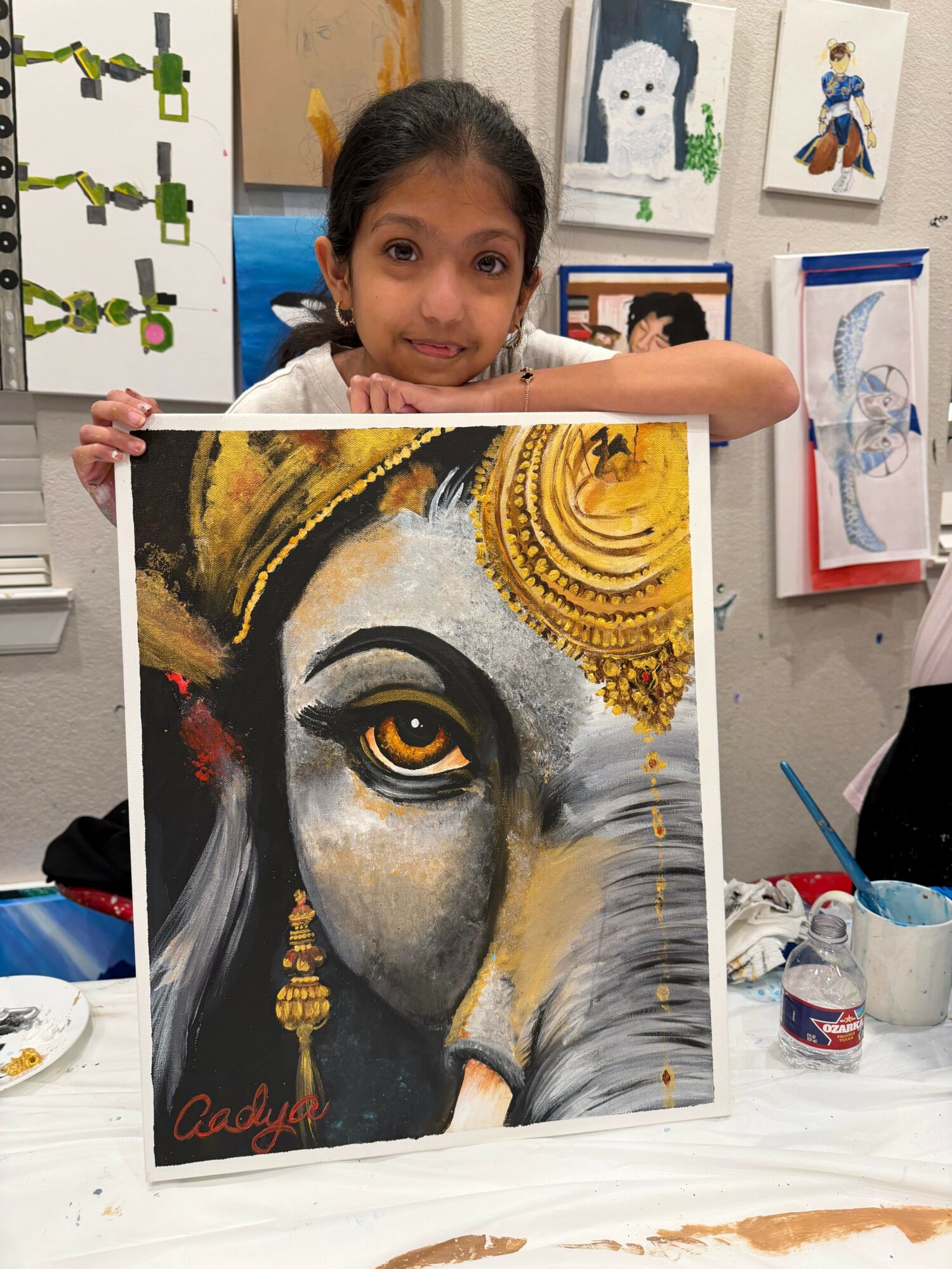 Young girl holding a painting of a woman's face with gold accents, sitting at a table with art supplies.