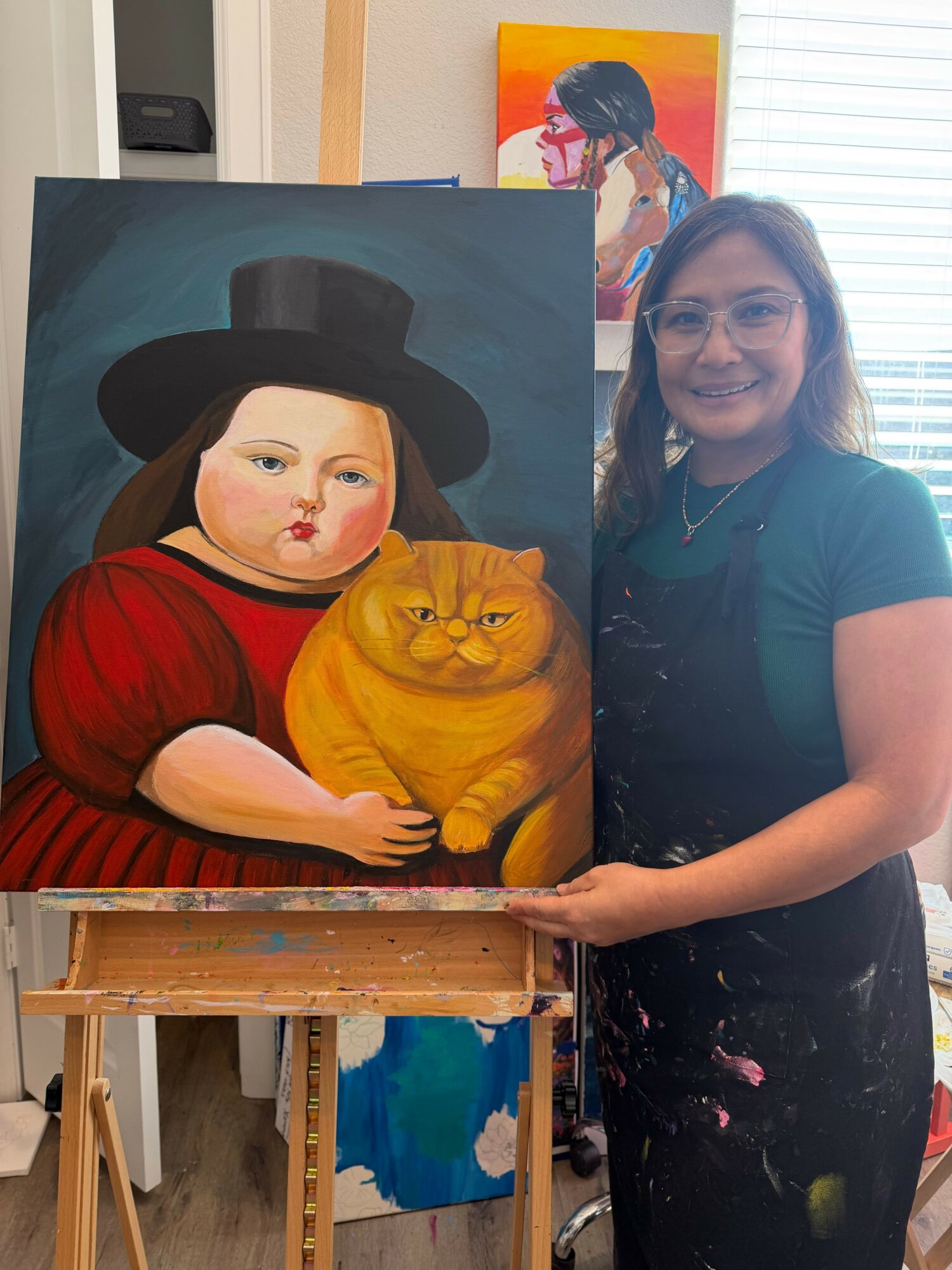 Artist smiling in studio, standing next to a colorful painting of a woman with a pumpkin, wearing a black hat and red dress.