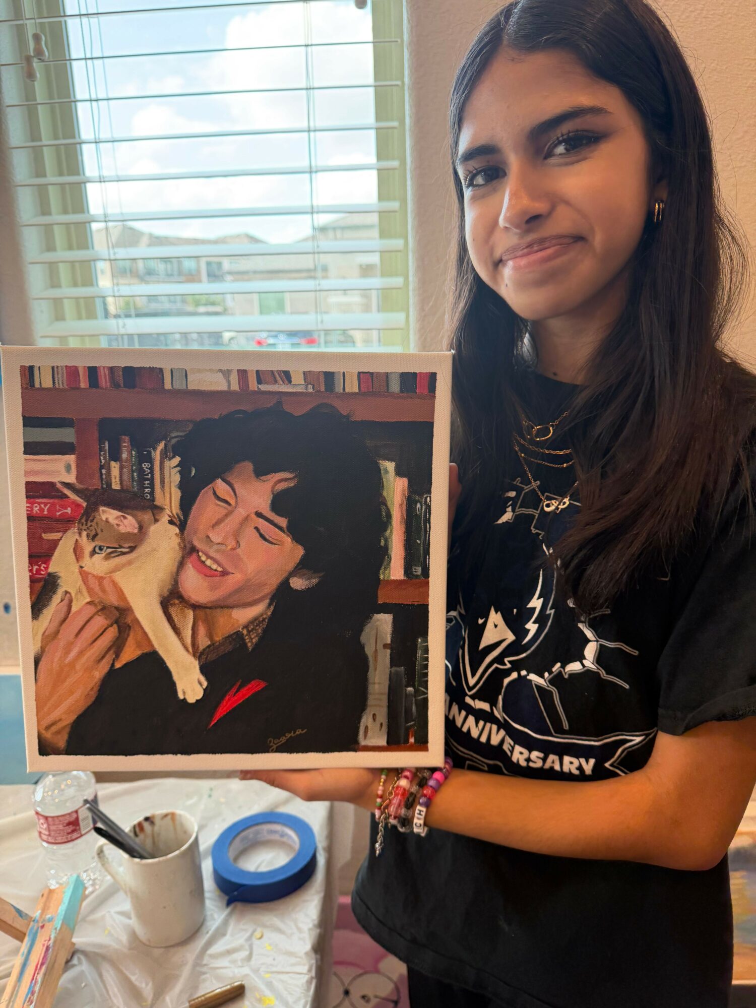 Person with long dark hair holding a framed photo of a woman with short dark hair smiling, in front of a window with blinds