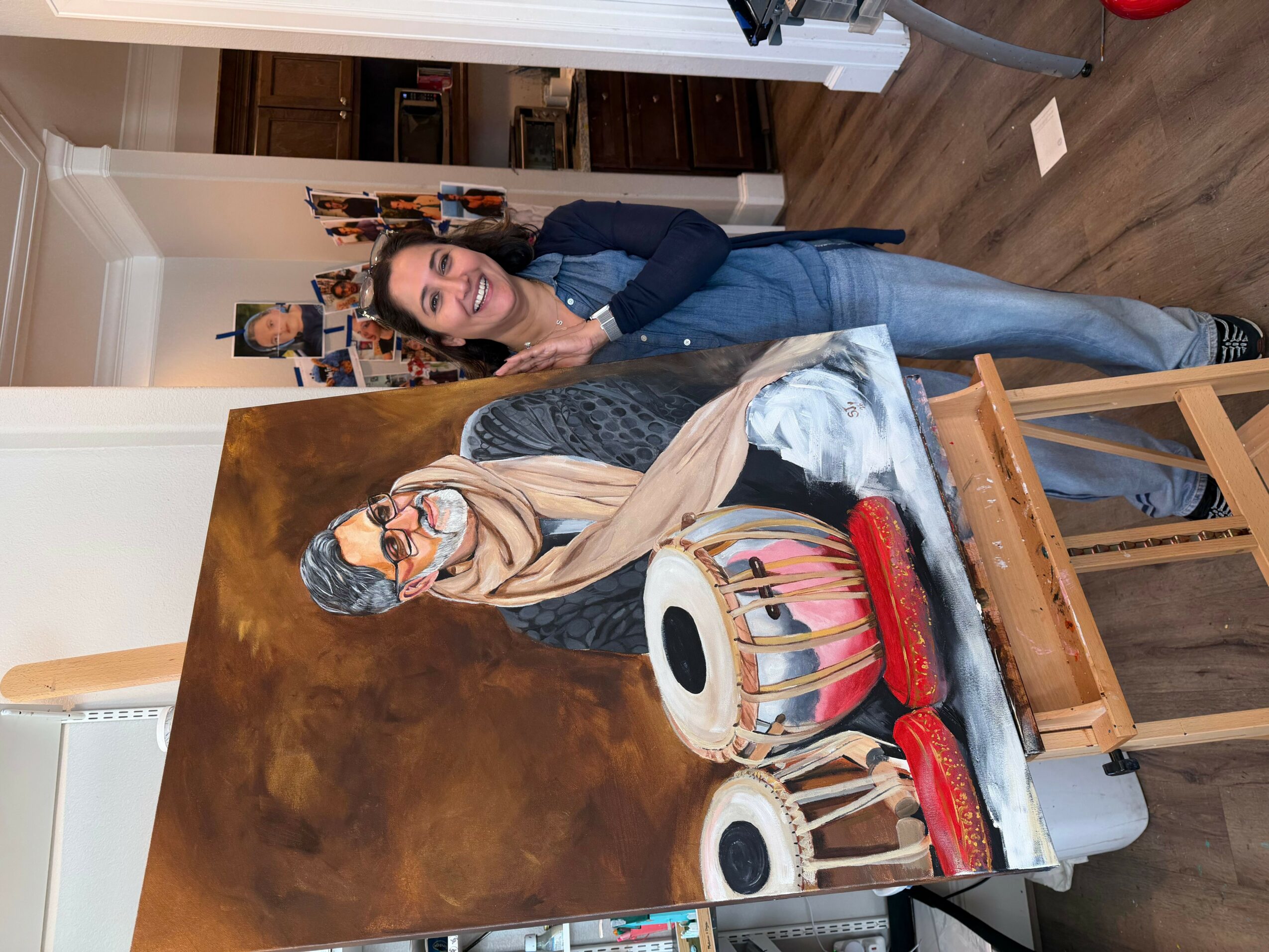 Woman with dark hair and glasses leaning over a painting of a woman with a headscarf and glasses, playing drums, in a room.
