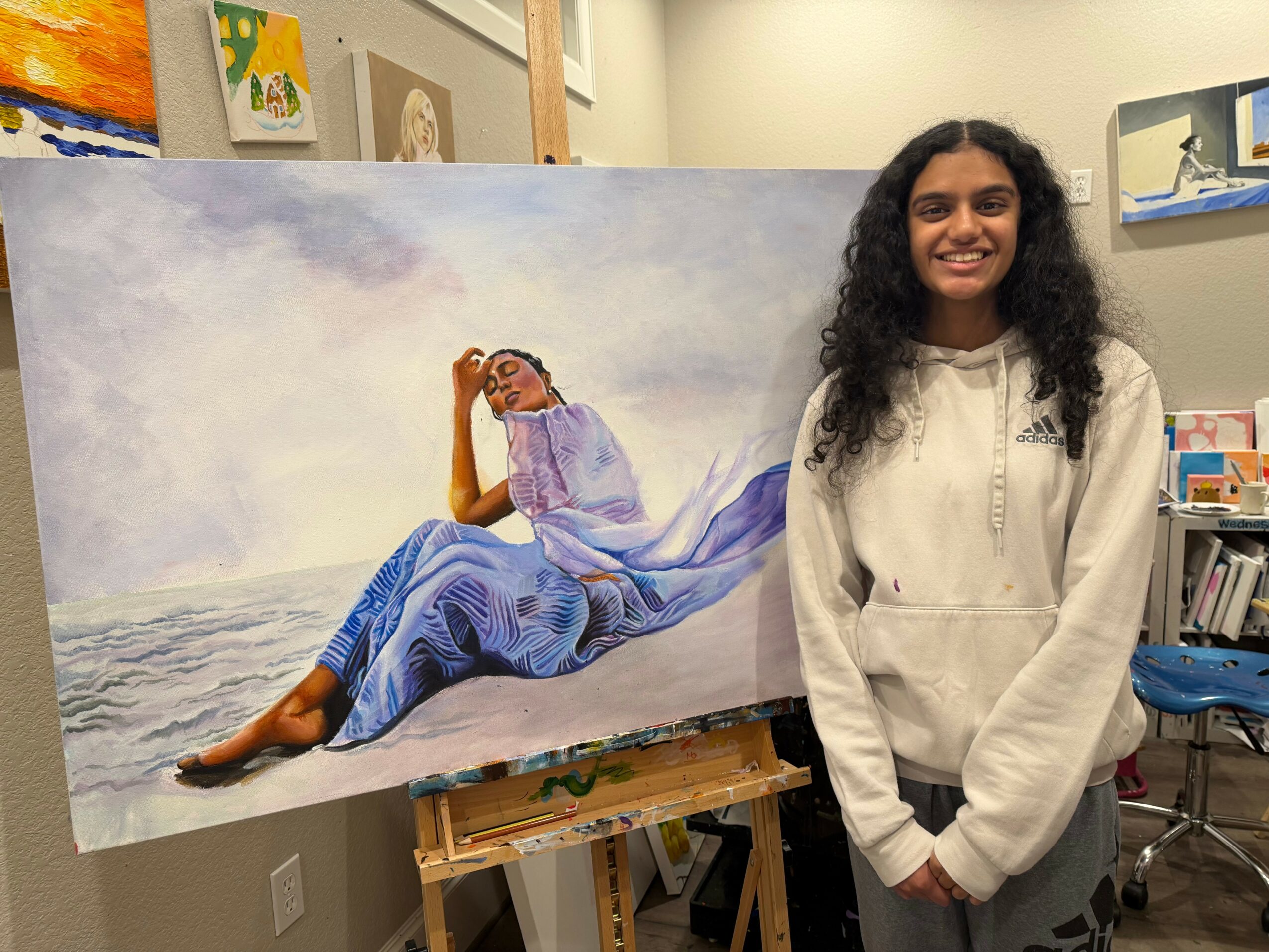 Young woman with curly hair smiling, standing in front of a large painting of a woman relaxing on a beach, wearing a purple dress.
