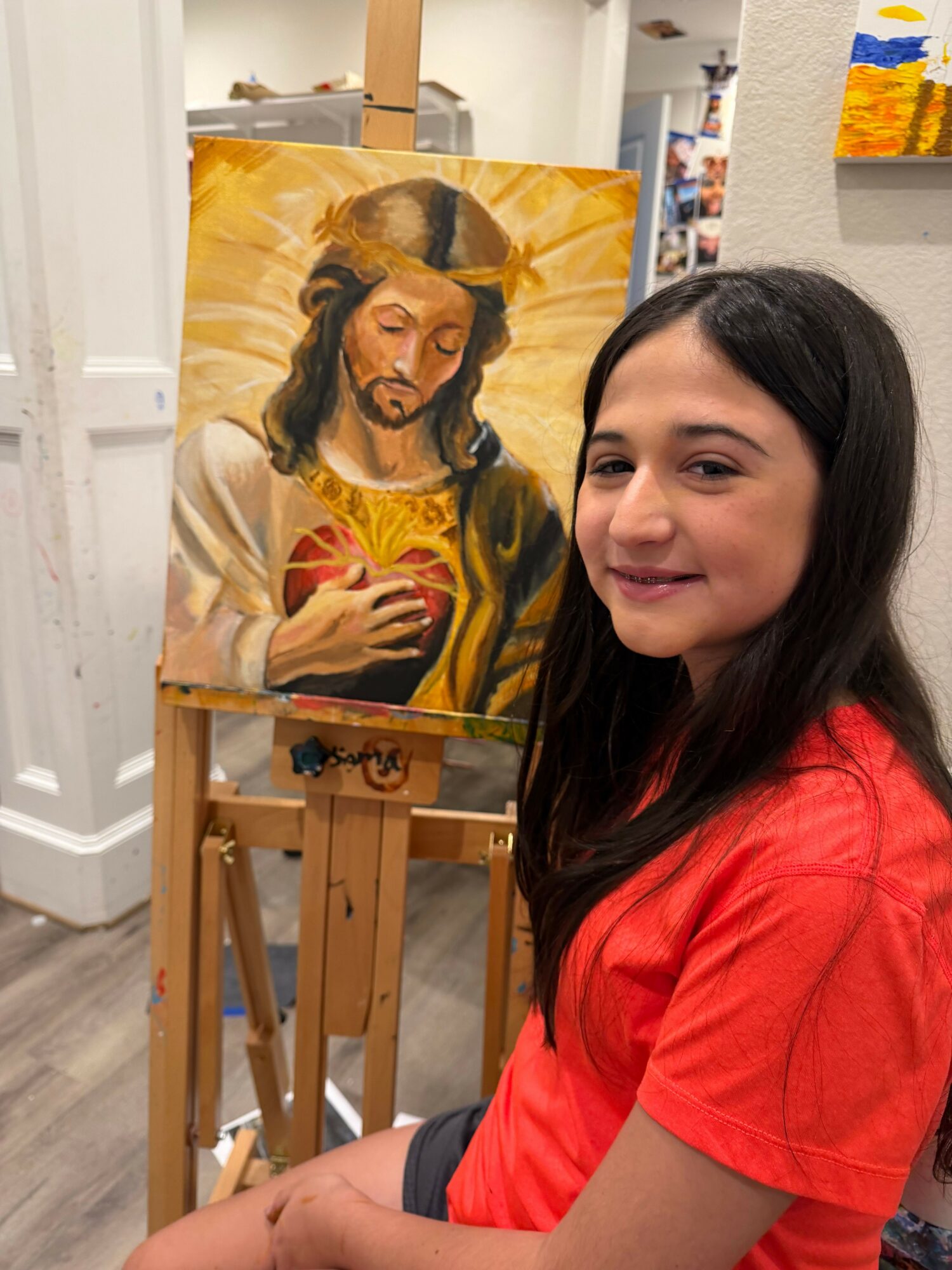 Young woman with long dark hair in a red shirt, smiling, sitting next to a painting of a woman holding a phone.