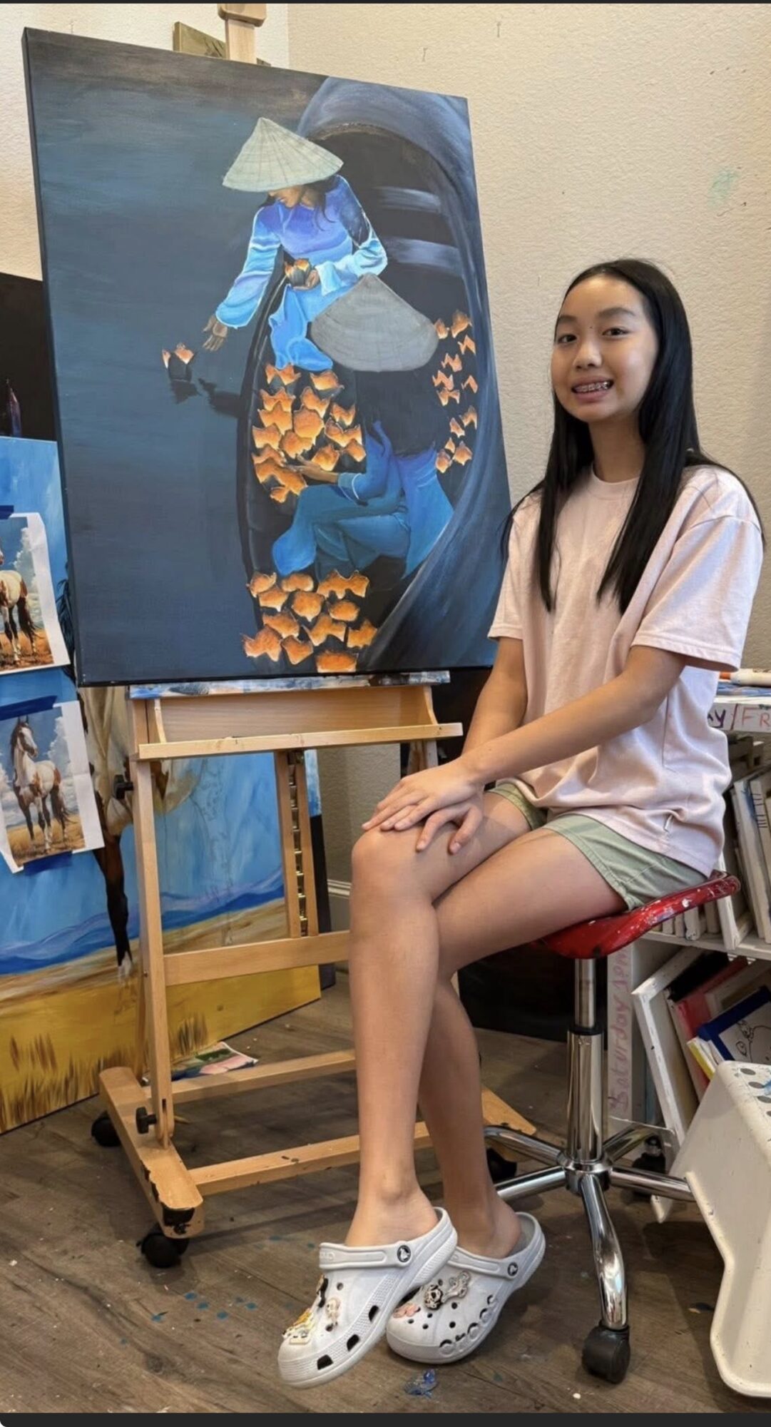 Young woman sitting on a stool in front of a large painting of a person in traditional attire, smiling.