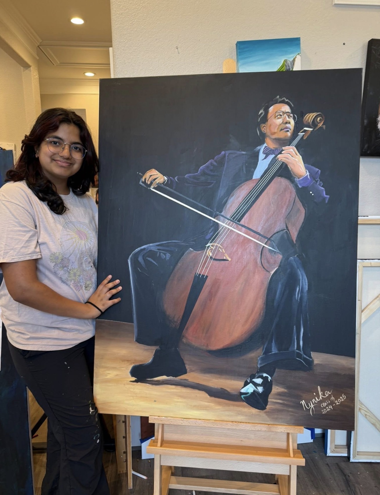 Young girl with dark hair and glasses standing next to a painting of a man playing a cello, smiling.