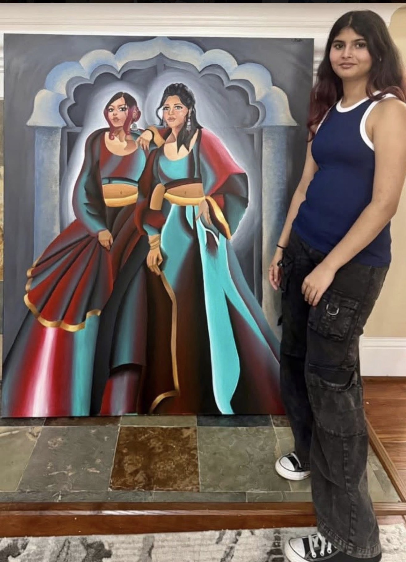 Young woman with long dark hair wearing a blue and white sleeveless top and black jeans standing next to a colorful painting of two women in traditional attire.