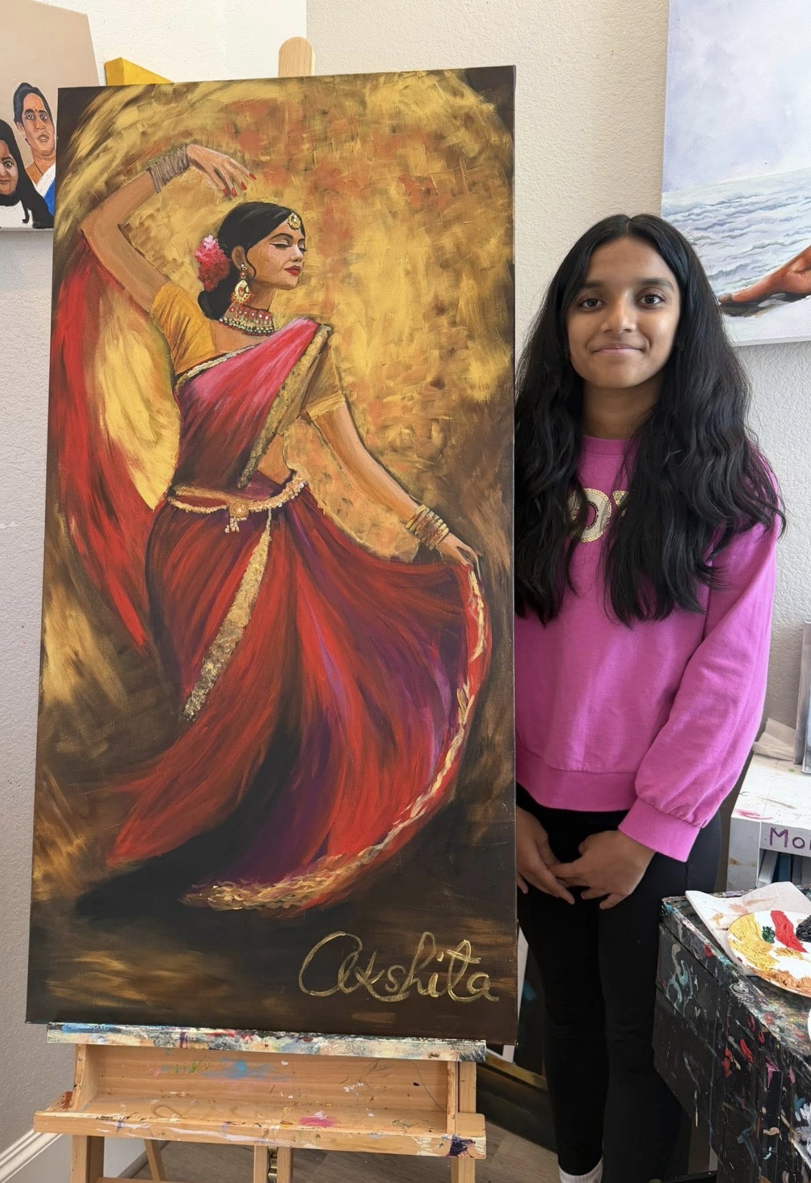 Young woman with long black hair in pink sweater standing next to a colorful dance painting on an easel.