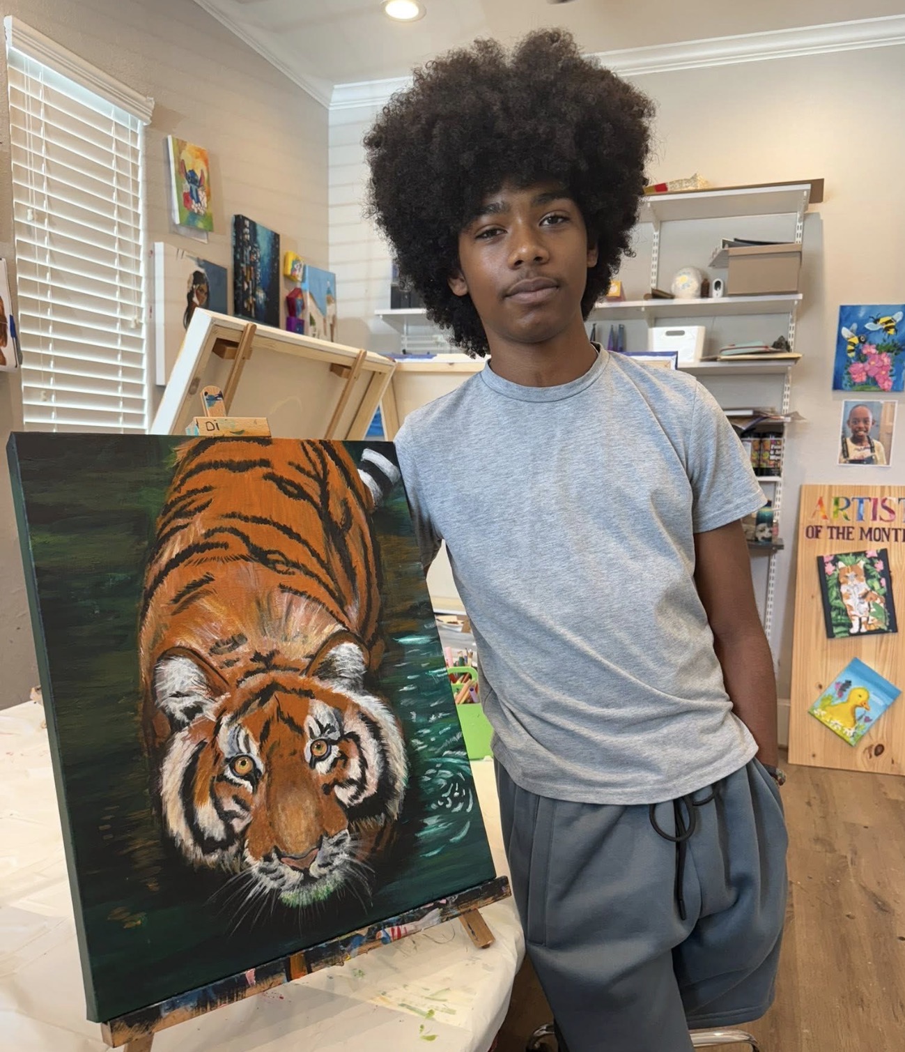 Young person with large afro hairstyle standing next to a tiger painting in a room with shelves and artwork.