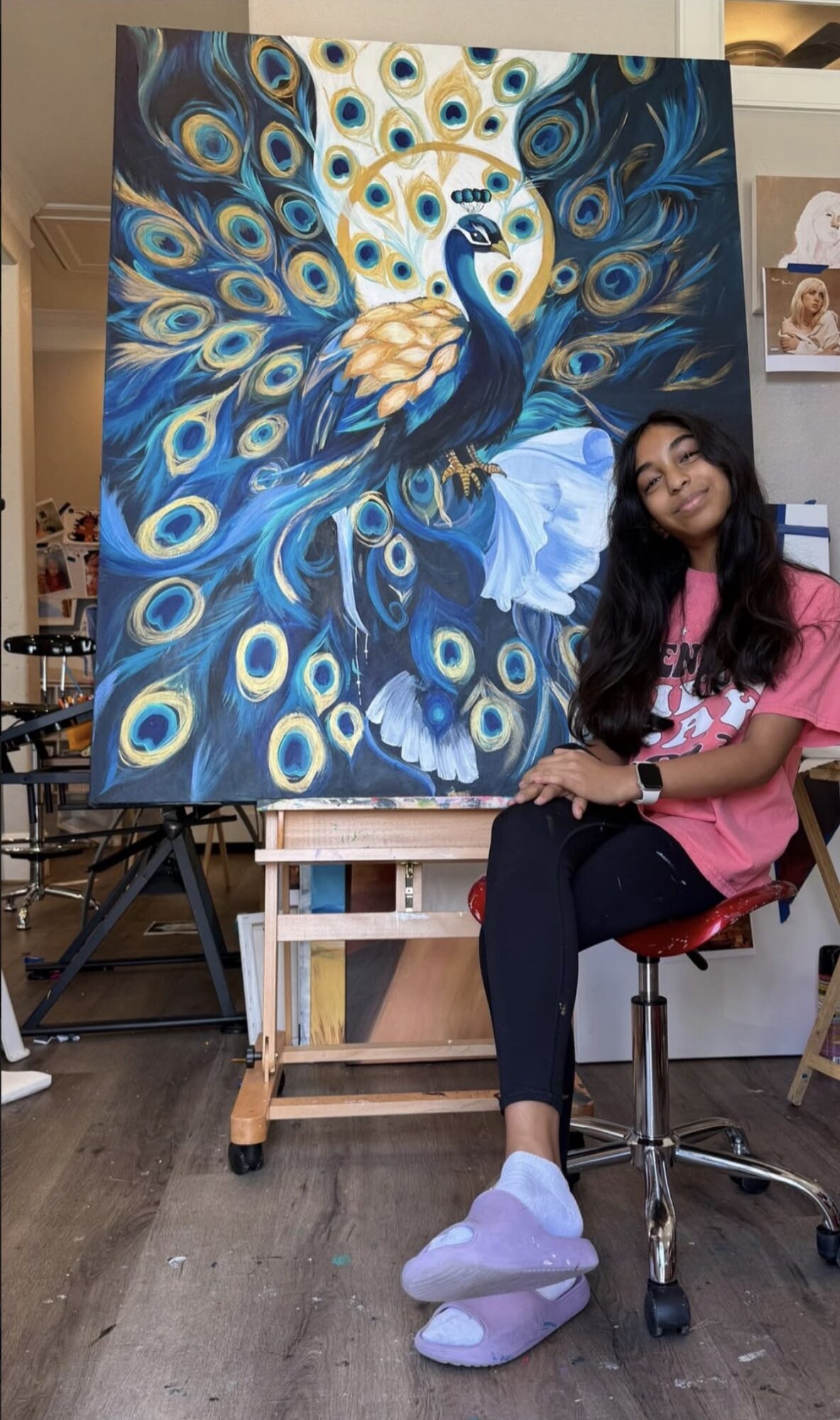 Young woman sitting in front of a large peacock painting with vibrant blue and gold feathers, smiling at the camera.
