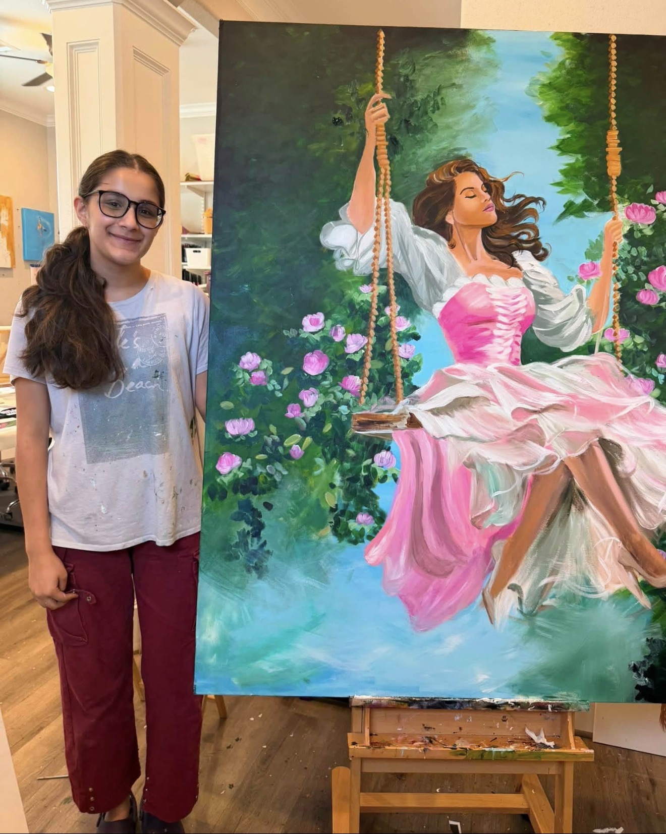Girl with glasses and long hair standing next to a colorful painting of a woman on a swing in a garden.