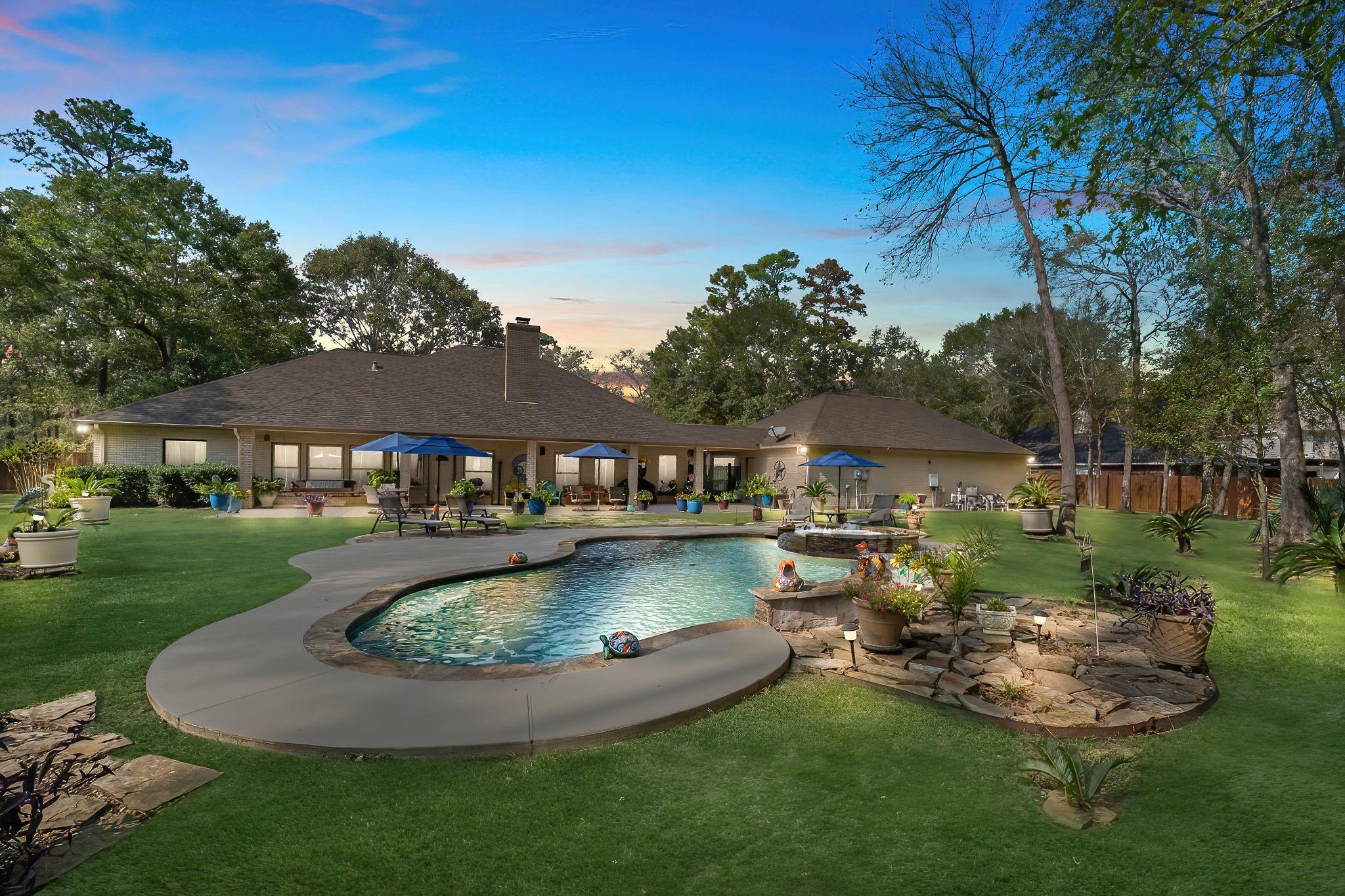 Backyard with swimming pool, patio, umbrellas, trees, and a house in the background under a blue sky.