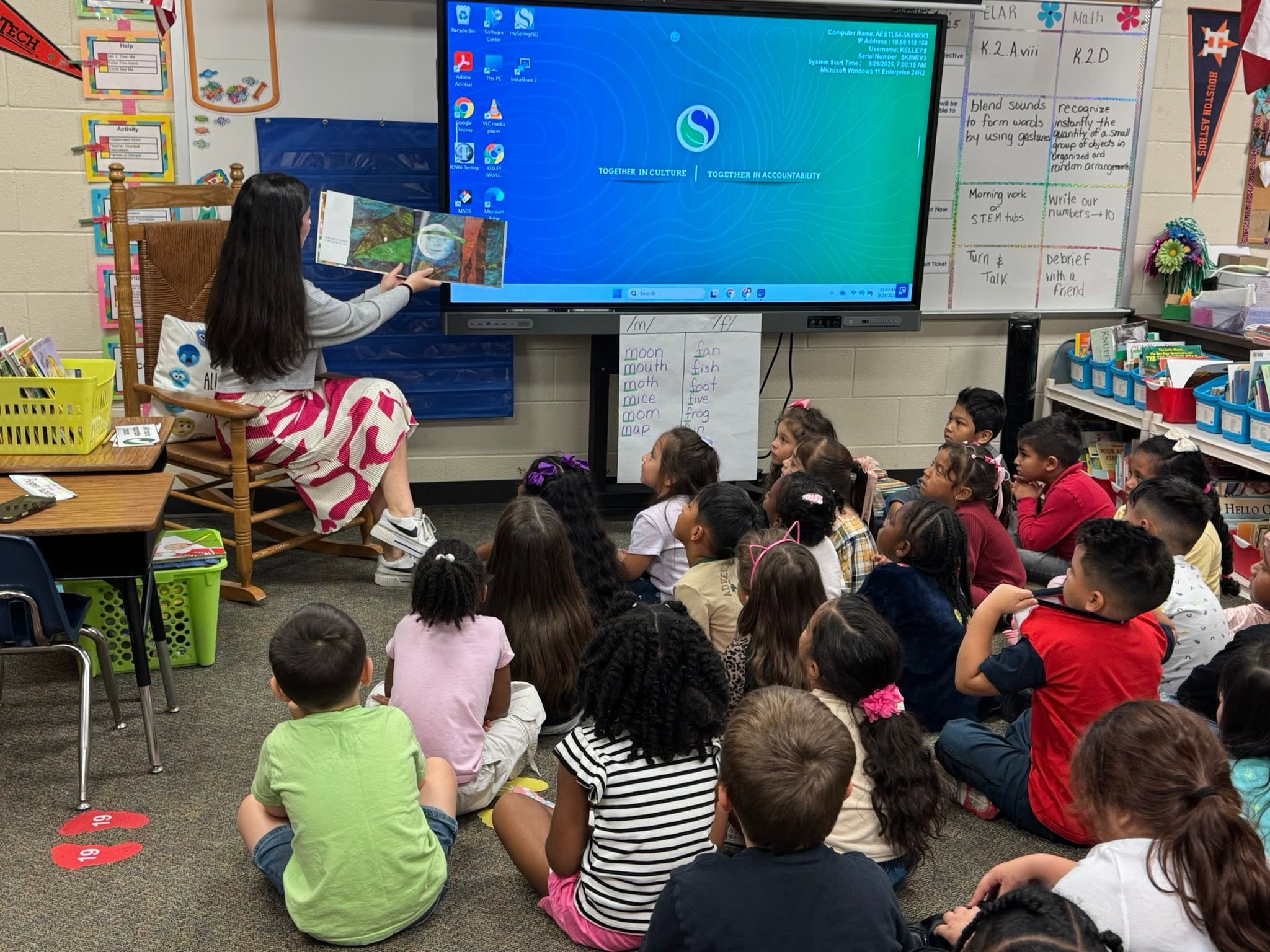 Teacher reading a book to children seated on the floor in a classroom with educational posters and a large screen.