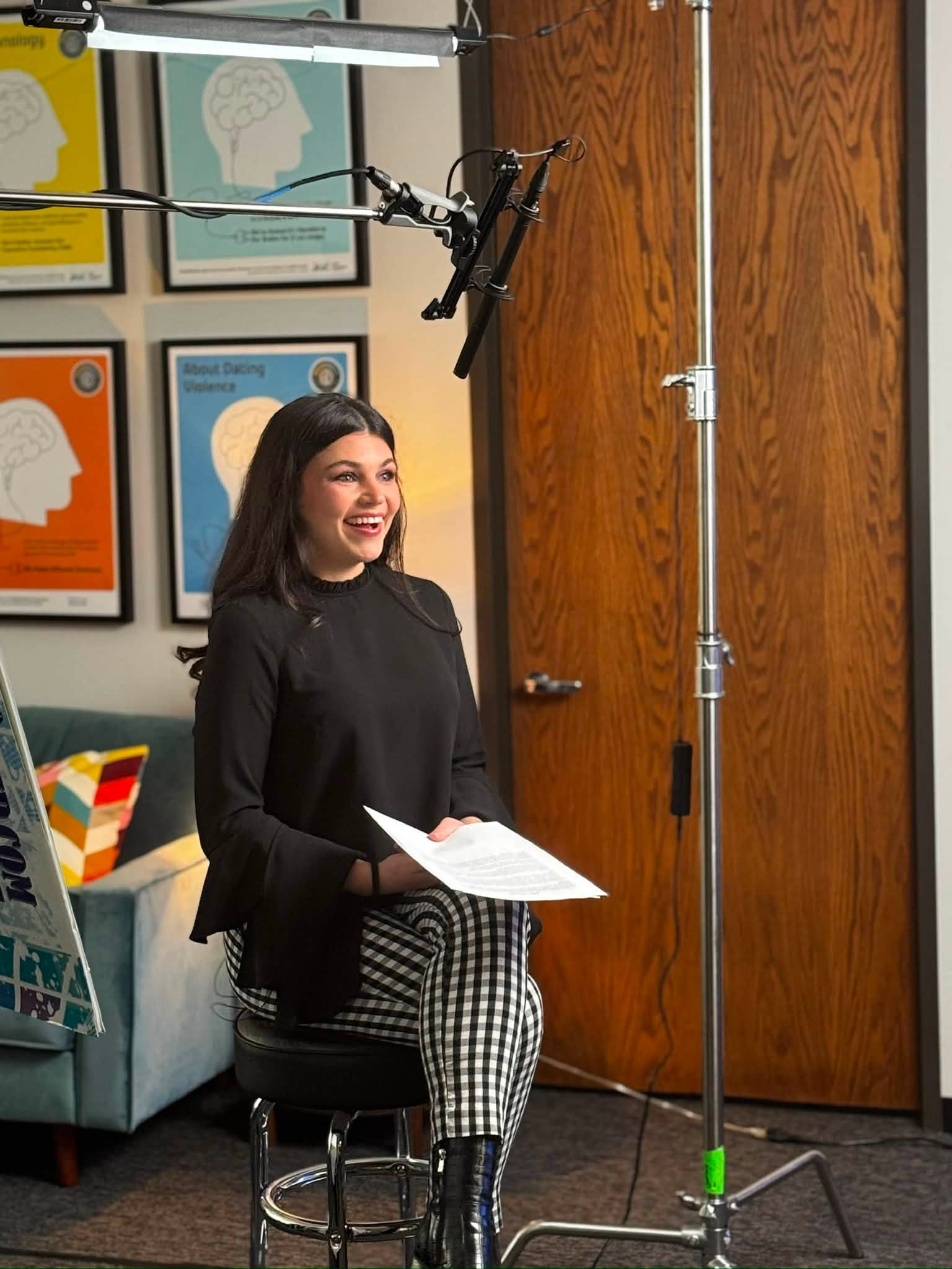 Woman sitting on a stool holding papers, smiling, in a room with framed artwork on the wall.