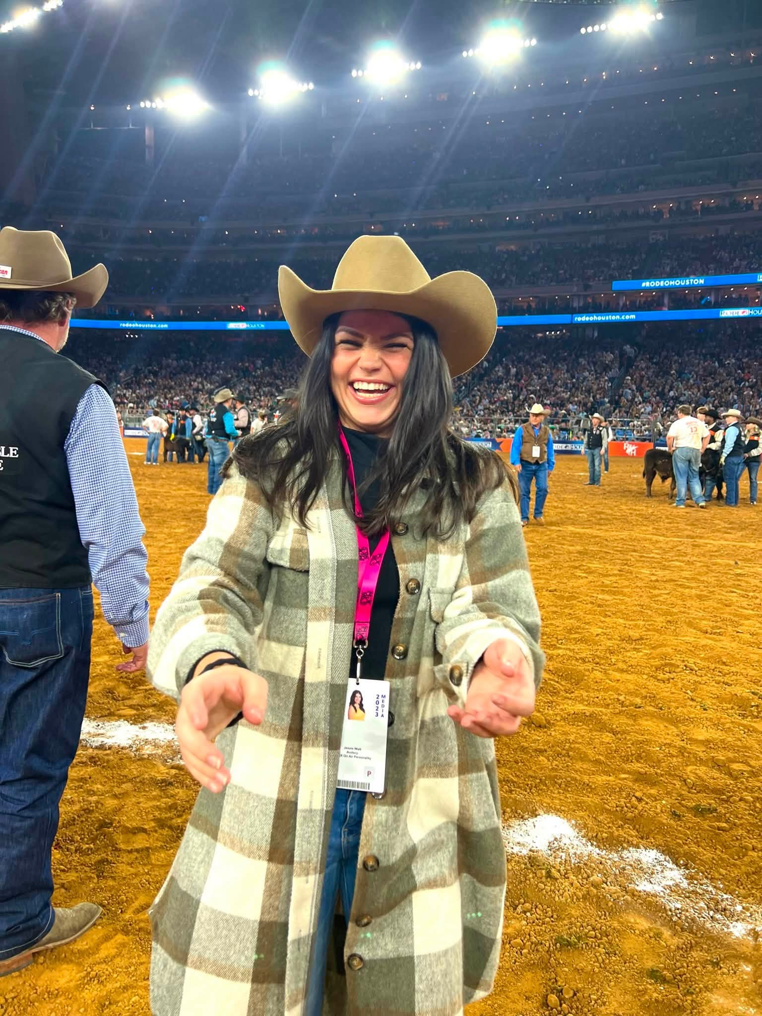 Smiling woman wearing a cowboy hat and plaid coat at a rodeo arena, with people and bright lights in background.
