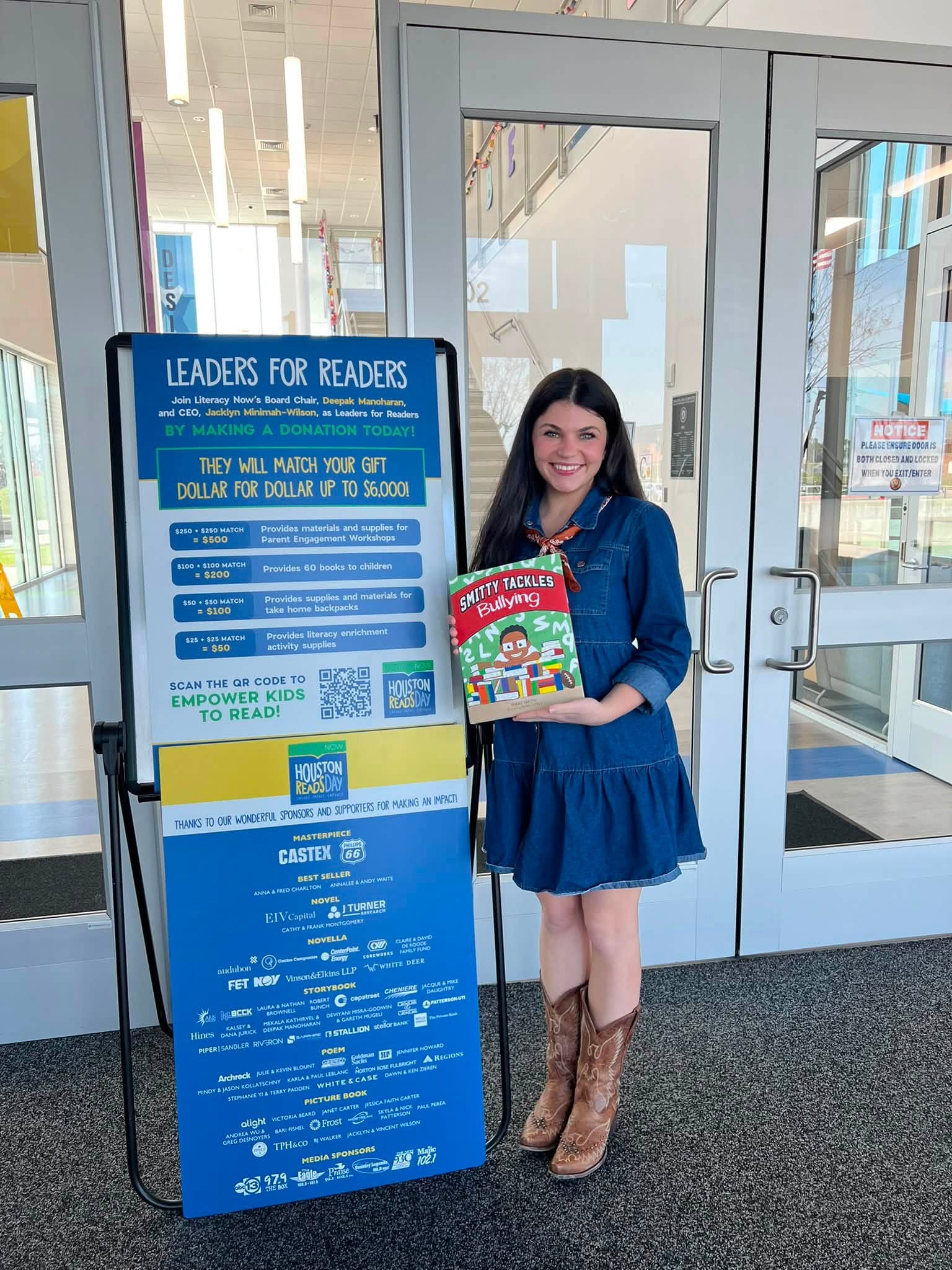 Young woman with dark hair, wearing a blue dress and cowboy boots, holding a colorful book, standing next to a blue informational sign inside a building.